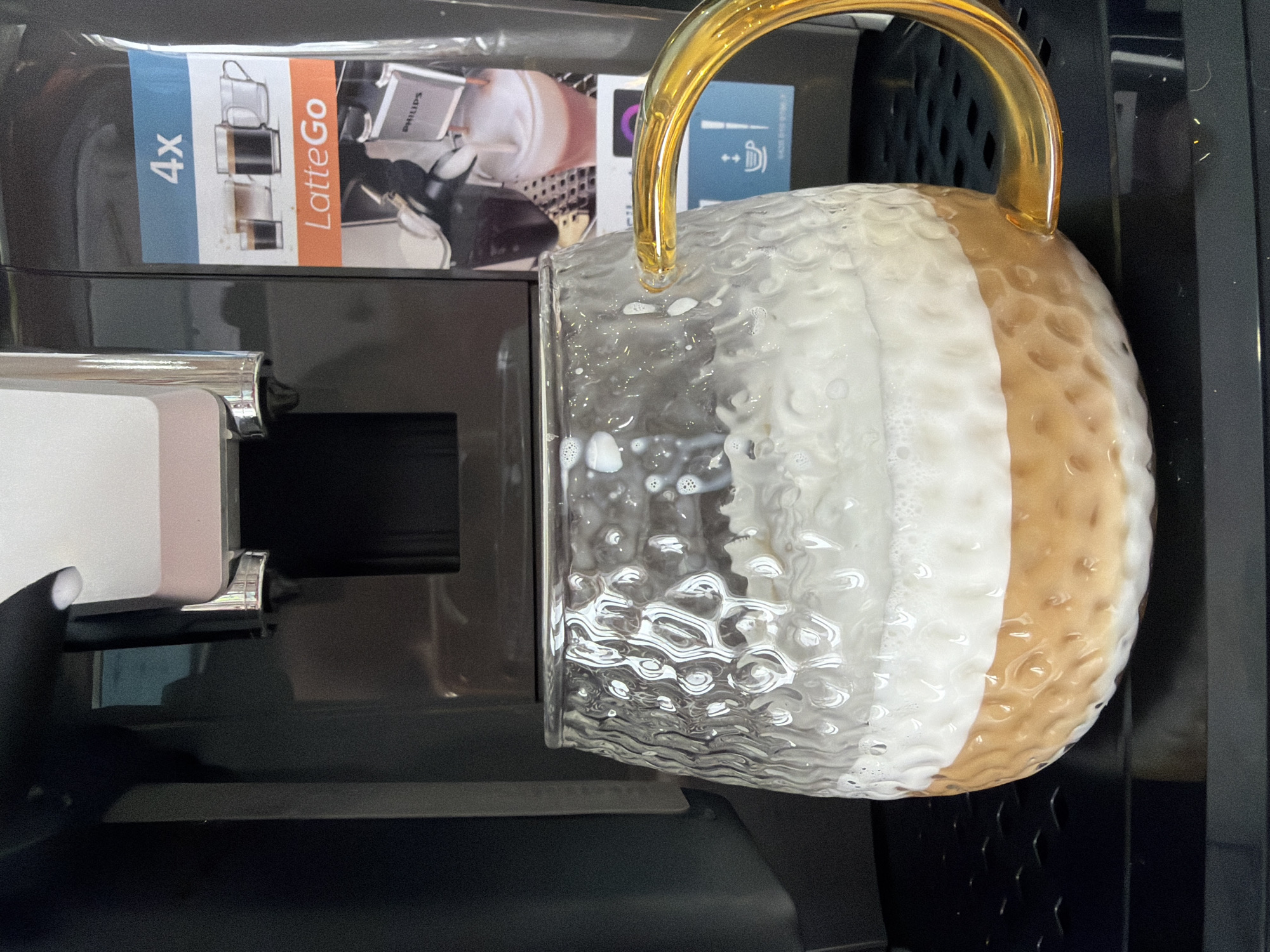 A glass mug with a golden handle and a two-toned drink (white and tan) is placed under the dispenser of a Philips LatteGo coffee machine. The machine is mostly black. There is a LatteGo packaging box partially visible. The photo was likely taken indoors. The location of Chaikovsky, Russia is not evident from this image.
