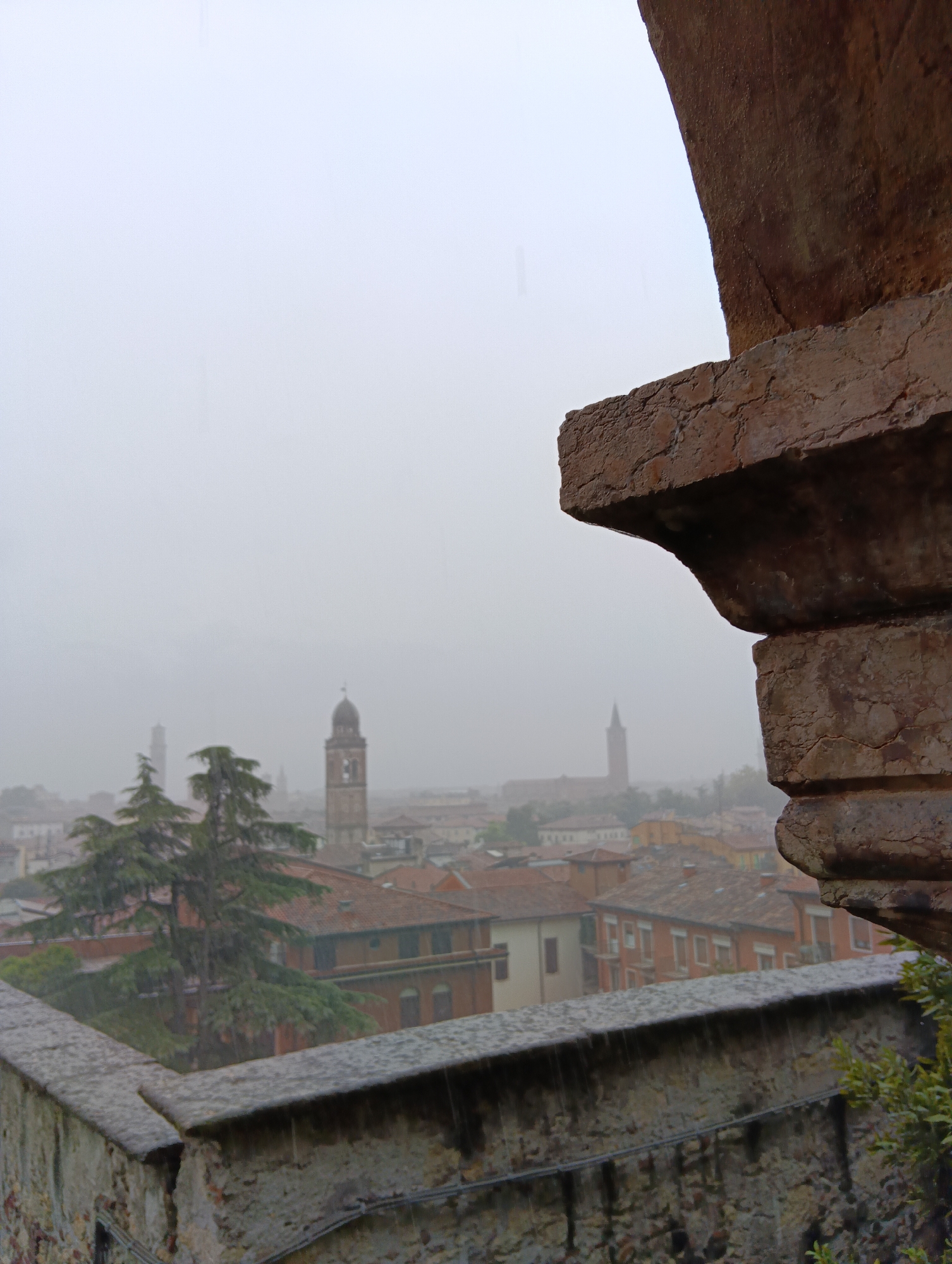 The image shows a view of Verona, Italy on a rainy day. In the foreground, a stone wall and a large, weathered stone structure are visible on the right side, framing the scene. Beyond the wall, a dense cluster of terracotta-roofed buildings stretches across the middle ground. Several church towers rise above the rooftops. The prominent tower on the right is characterized by its brick construction and a dark, dome-like spire. Another, taller tower is visible further in the distance, appearing more slender and elongated due to the atmospheric haze. A tall, dark green coniferous tree stands to the left of the main cluster of buildings. The sky is overcast and a light rain is falling, indicated by faint streaks in the air. The overall atmosphere is muted and hazy, typical of rainy weather.