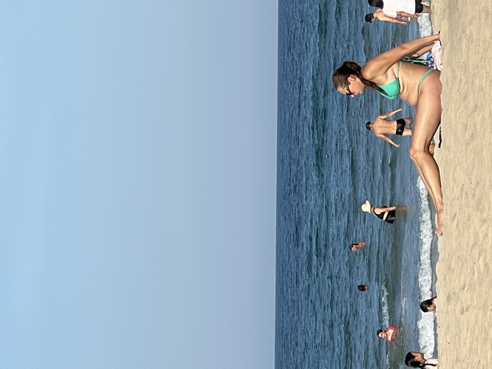 In Đà Nẵng, Vietnam, this image depicts a sunny day at the beach. A woman in a teal bikini sits on the sand in the foreground, facing right, her legs extended towards the ocean. Behind her, several people are in the water. A man in dark swim trunks stands with his back to the camera. Further out, a woman in a black swimsuit and a wide-brimmed hat is visible, as are others swimming. The sea is a deep blue, with small waves breaking near the shore. The sky above is a clear, pale blue.