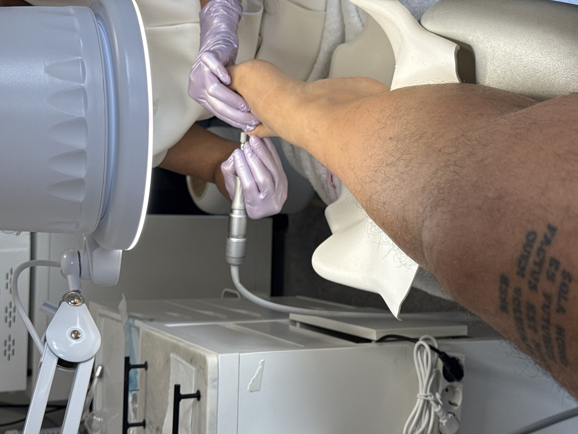 A foot care procedure is being performed, possibly a pedicure, on a person with a tattoo on their lower leg. The tattoo is in a vertical orientation and appears to be Latin script. A technician wearing lavender-colored gloves is using a handheld rotary tool on the foot. The foot rests on a white surface or chair, part of which is visible in the upper right. A white articulated lamp is positioned to the left, illuminating the foot. The scene takes place indoors in what appears to be a salon or treatment room, with white cabinets and equipment in the background. There is no available information regarding the city of Differdange, Luxembourg.