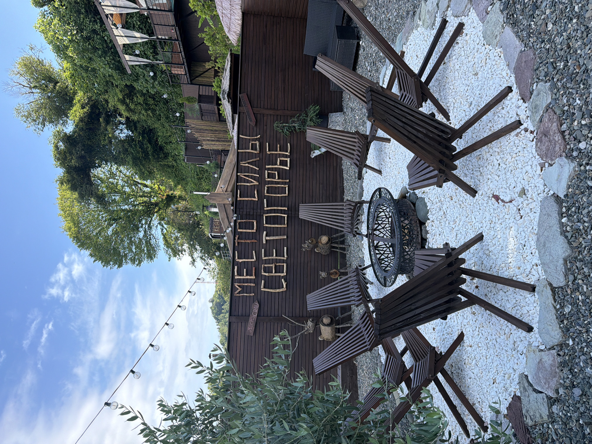 The image depicts an outdoor seating area with a fire pit, located in Nizhnyaya Shilovka, Russia. Dark wooden chairs are arranged in a circle around a metal fire pit, which sits on a bed of white pebbles. Behind the chairs, a dark wooden fence displays large letters made of branches that read "МЕСТО СИЛЫ СВЕТОГОРЬЕ" (Mesto Sily Svetogor'ye), translating to "Place of Power Svetogor'ye." Smaller wooden signs are also visible on the fence. The scene is surrounded by lush green foliage and trees, with a multi-tiered wooden structure and thatched roofs visible in the background, suggesting a resort or recreational area. String lights are suspended overhead, and the sky is partly cloudy. No people are present in the image.