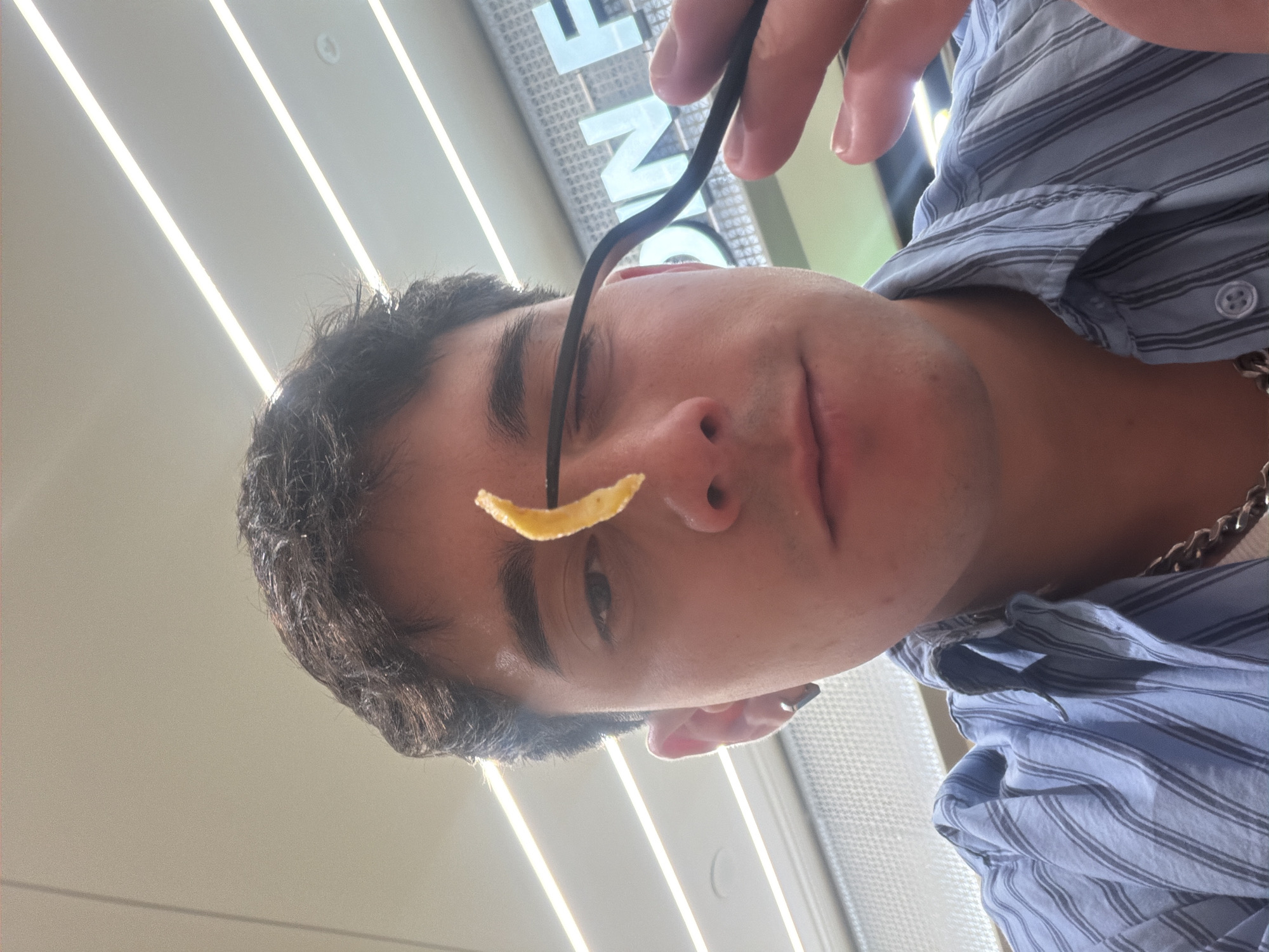 A young man is holding a french fry on a fork in front of his face. He is wearing a blue striped button-down shirt and a silver chain. The location appears to be indoors under fluorescent lighting, possibly in a food court or restaurant. While it's challenging to definitively confirm the location in Sofia, Bulgaria, without more context, the fluorescent lighting and potential signage in the background suggest an indoor space where food is served.