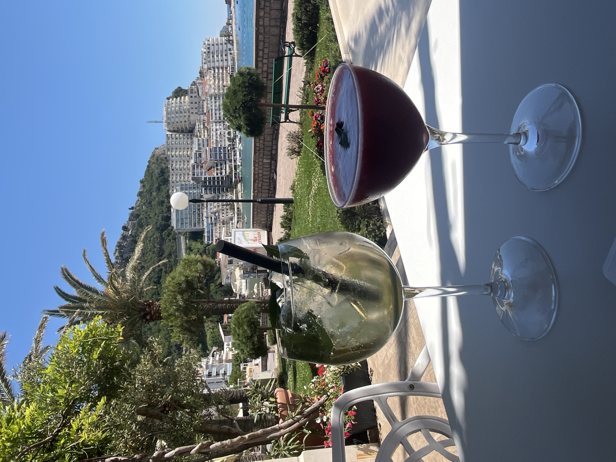 The image shows two cocktails on a white table, set against a view of Bečići, Montenegro. The cocktail closest to the viewer is a light-colored beverage with ice and a black straw in a large glass. The second drink is a darker red color, also in a large glass. Behind the cocktails is a view of the city with buildings and mountains in the background. A palm tree and other greenery are visible in the foreground, partially obscuring the view of the city. The weather appears sunny and clear.