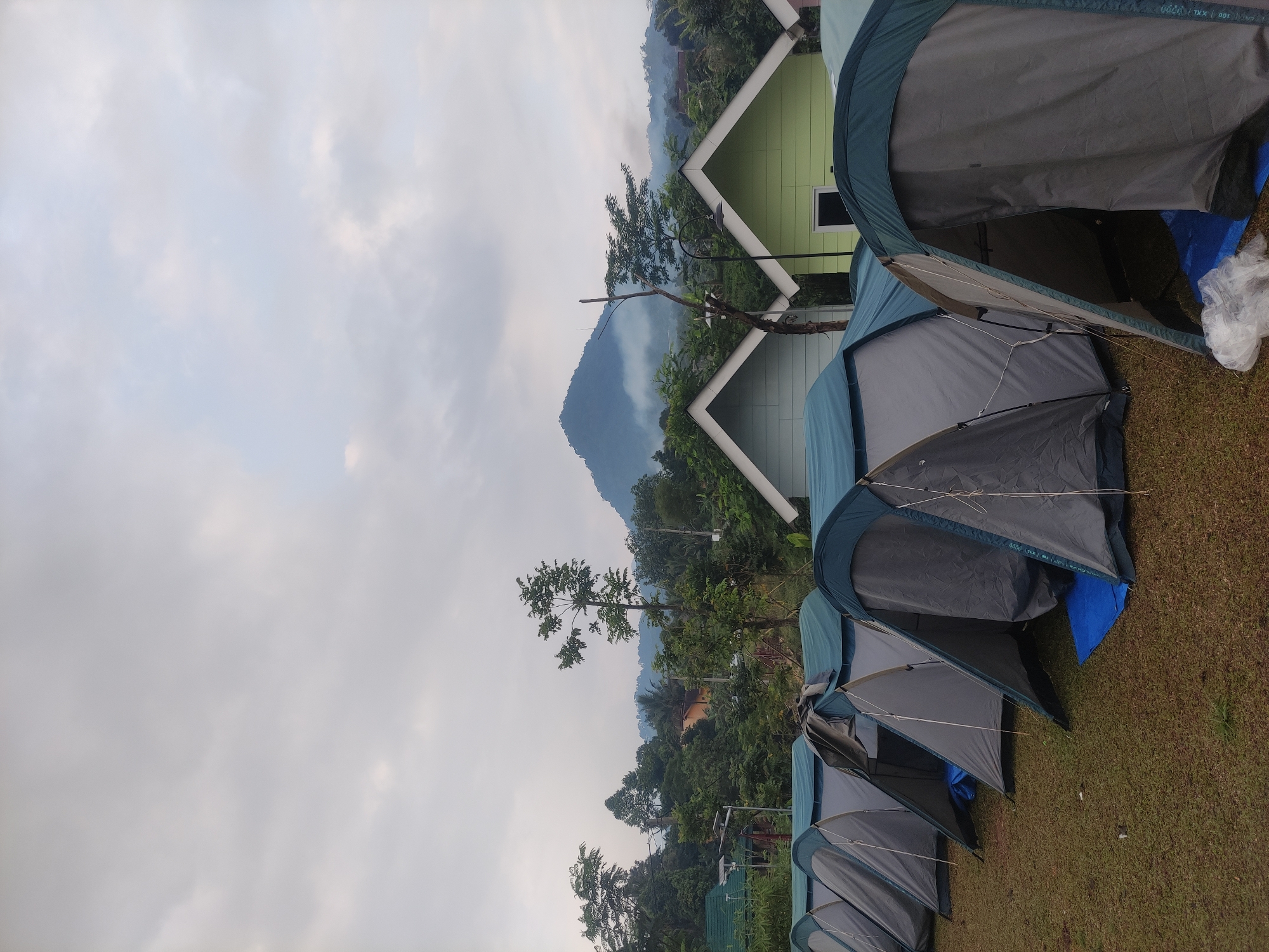 The scene depicts a campsite in Babakan Madang, Indonesia. Several grey and blue tents are pitched on a grassy field, with the front flaps open. In the background, a light green, gabled-roof building stands adjacent to the tents, with trees and a mountain visible behind it. The sky is overcast.
