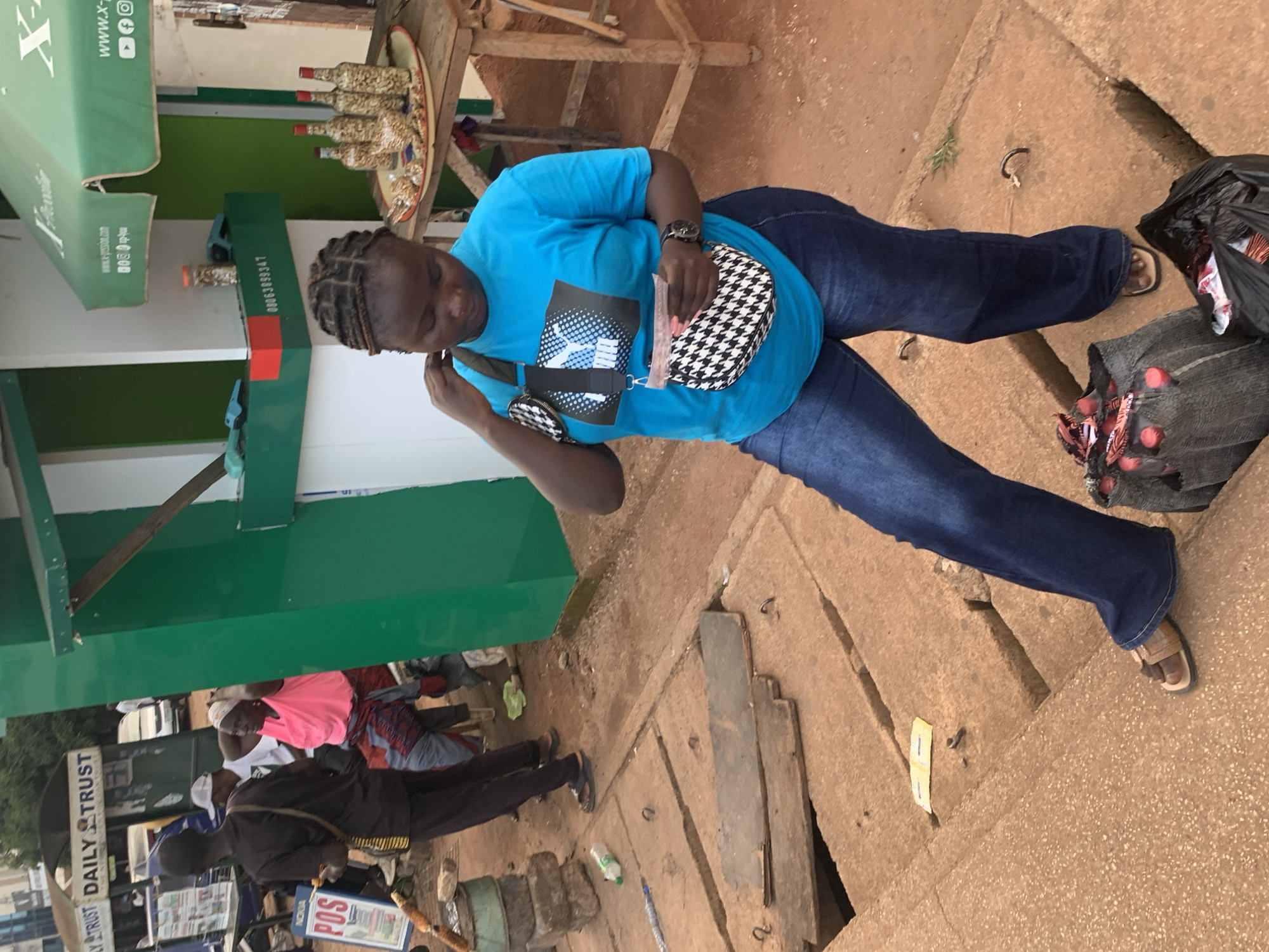A woman in a bright blue t-shirt and blue jeans stands on a concrete surface, looking down at her hands. She has dark skin and her hair is styled in braids. She is holding a small purse and what appears to be a piece of paper or money. Her left hand is touching her hair. To her left, a man in a black shirt and pants walks away from the camera, carrying a stick and a bag. Behind him, another man wearing a pink shirt and a patterned skirt sits down. In the background, there is a green and white kiosk with a sign that reads "DAILY TRUST". There are also bottles and other items on a wooden table to the right. The scene appears to be an outdoor market or street in Jos, Nigeria.