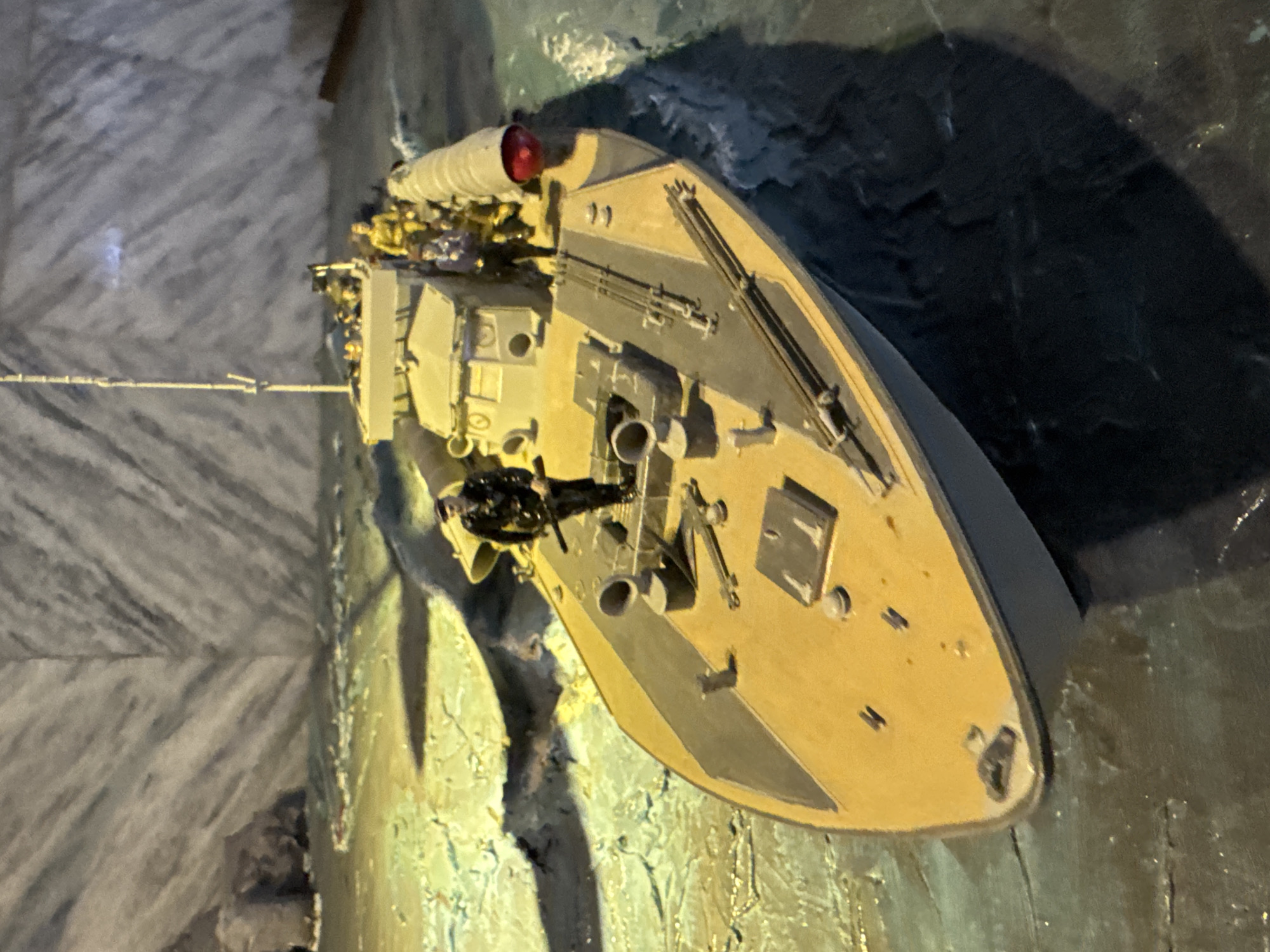 The image shows a model of the armored gunboat "Kanonerka" (Gunboat) on display in Saint Petersburg, Russia. The model is situated against a backdrop that simulates a rough sea or rocky terrain. It has a flat deck, light tan color, and various military-style details such as a machine gun, cylindrical tubes, and hatches. The shadows on the right suggest an artificial lighting source. The backdrop on the left appears to be a wall with a rough texture.