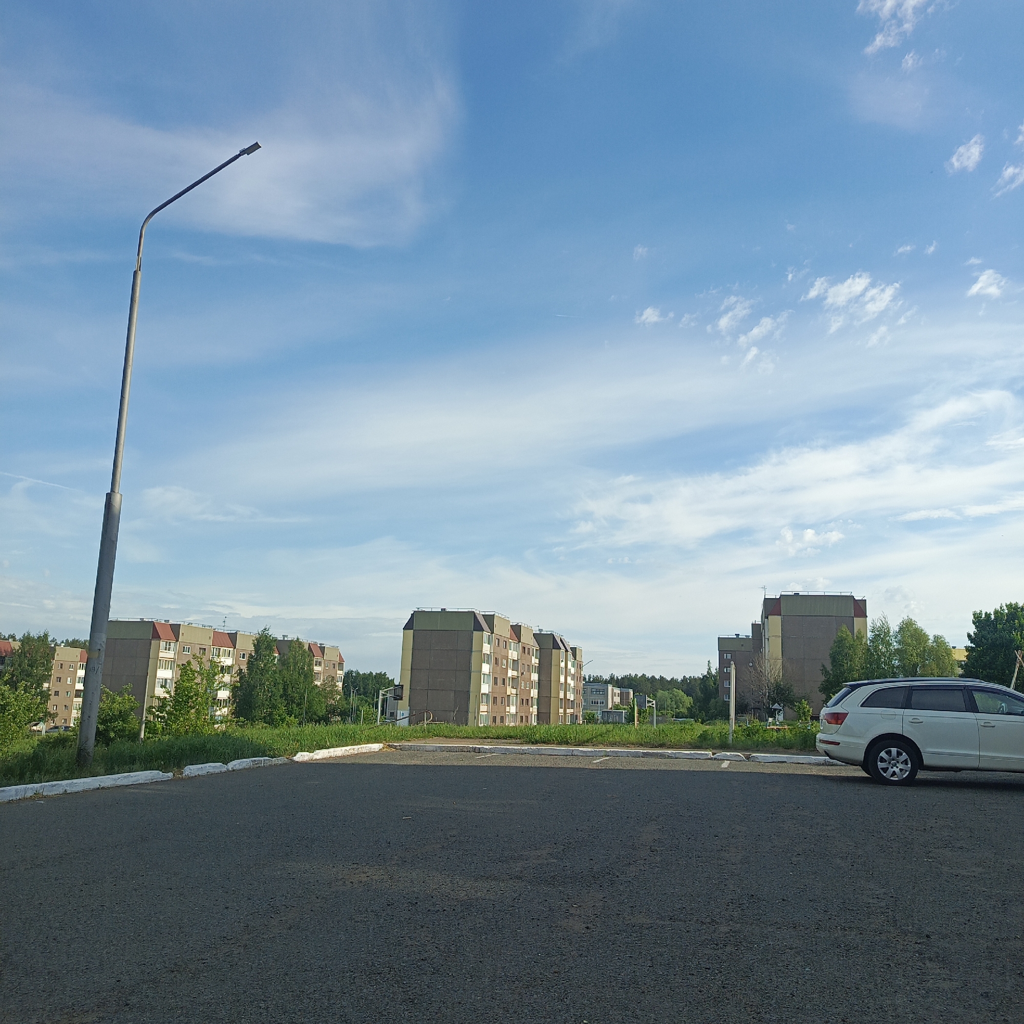 The image captures a cityscape in Марковский, Russia, featuring a parking area in the foreground. The asphalt pavement occupies the lower portion of the frame. In the background, a series of apartment buildings stand, characterized by their uniform, boxy design and a combination of grey and beige facades with red roofs. Green trees and foliage partially obscure the buildings, indicating some landscaping. A white SUV is parked on the right side of the frame. A tall lamppost is located on the left side of the frame. The sky above is blue with scattered white clouds.