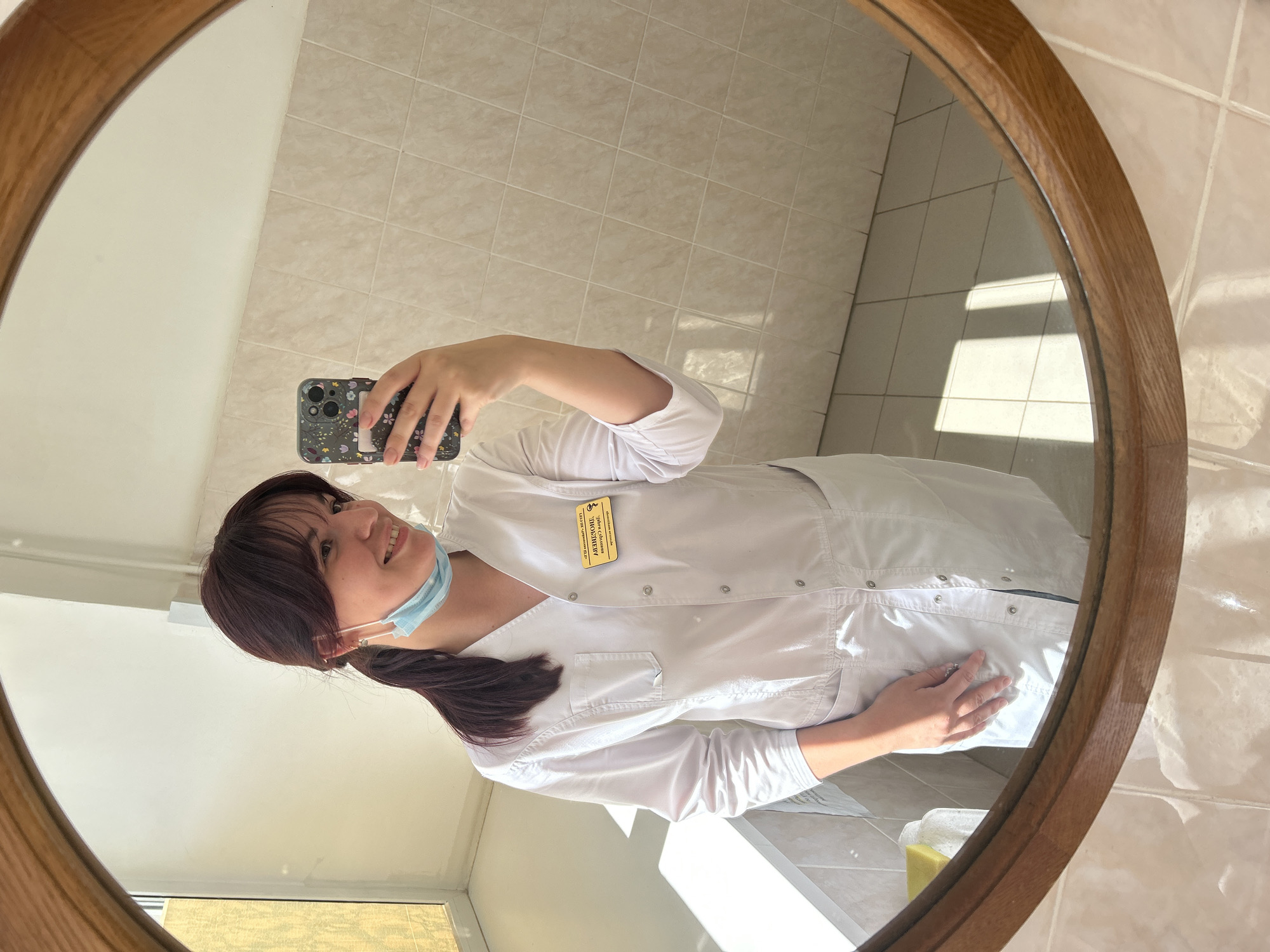 The image is a selfie taken in a mirror by a young woman. She is wearing a white medical coat, a light blue face mask, and has dark hair pulled back in a ponytail. A nametag is pinned to her coat, and while the text is partially obscured, it appears to be in Cyrillic script. She is smiling and looking at the camera. The background shows a tiled wall and floor, suggesting an indoor setting, possibly a bathroom or changing room. Sunlight is streaming in, creating distinct shadows on the tiled surface, indicating it is daytime. The mirror has a wooden frame. The overall impression is a casual, everyday moment captured in a reflective surface.