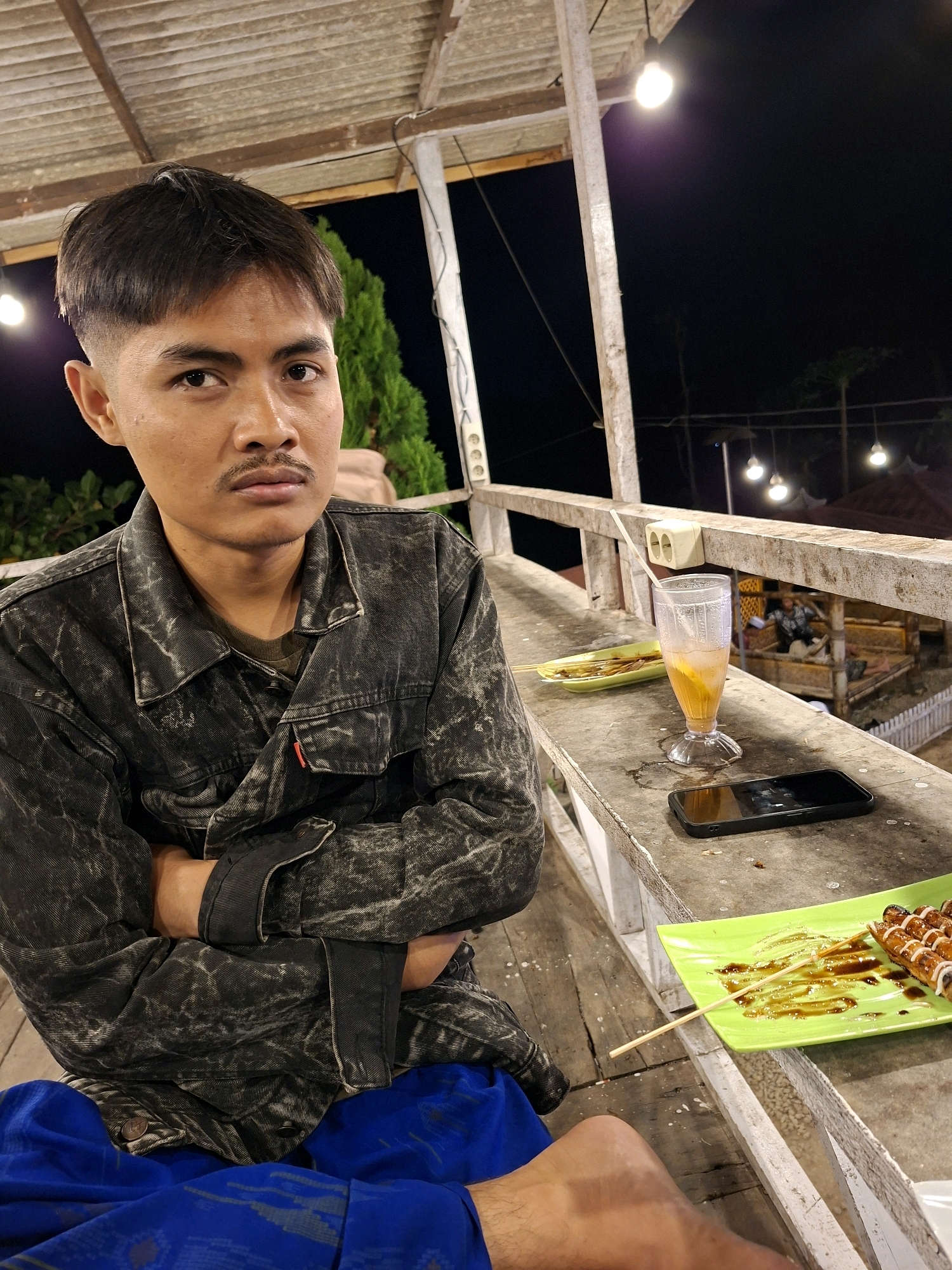 A young man sits at an outdoor table at night, with his arms crossed and a serious expression. He wears a dark denim jacket over a green t-shirt and blue pants. In front of him on the table are two green plates, one with skewers of grilled food drizzled in sauce and another with some sort of snack. A glass of orange liquid with ice and a straw is also on the table, as well as a smartphone. The setting appears to be a casual eatery or warung in Lantek Barat, Indonesia, characterized by wooden structures, open sides, and overhead lighting. In the background, other patrons can be seen in a gazebo-like structure. The overall atmosphere is dimly lit and relaxed.