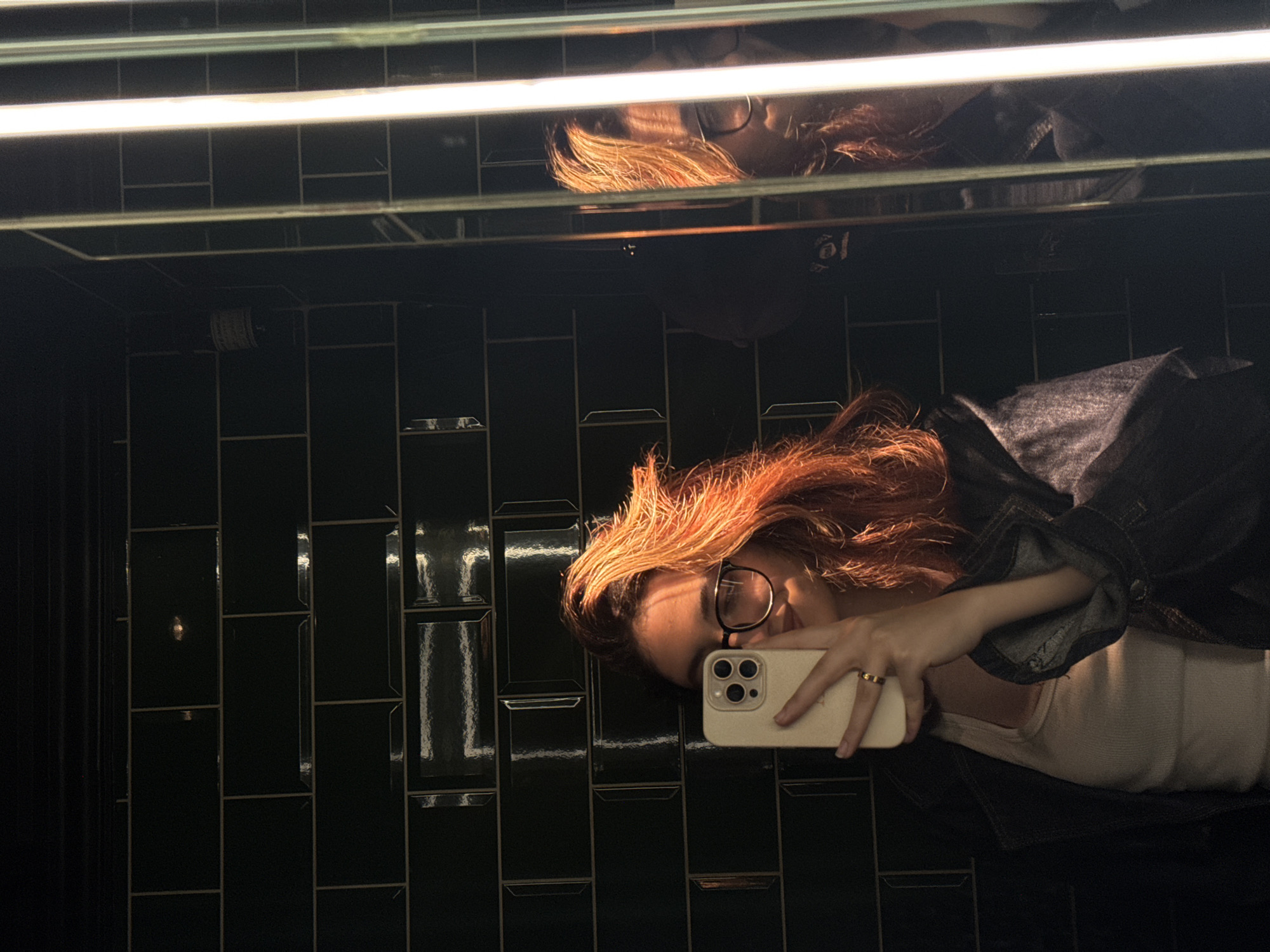A person with reddish-brown hair is taking a selfie in a restroom with dark green tile walls. The person is wearing glasses, a white shirt, and a denim jacket. They are holding a beige-colored smartphone in their right hand, capturing their reflection in a mirror above the sink. Two fluorescent lights are visible above the mirror. The background is dominated by the dark green tiled wall. There is no indication of this image being in Yerevan, Armenia.