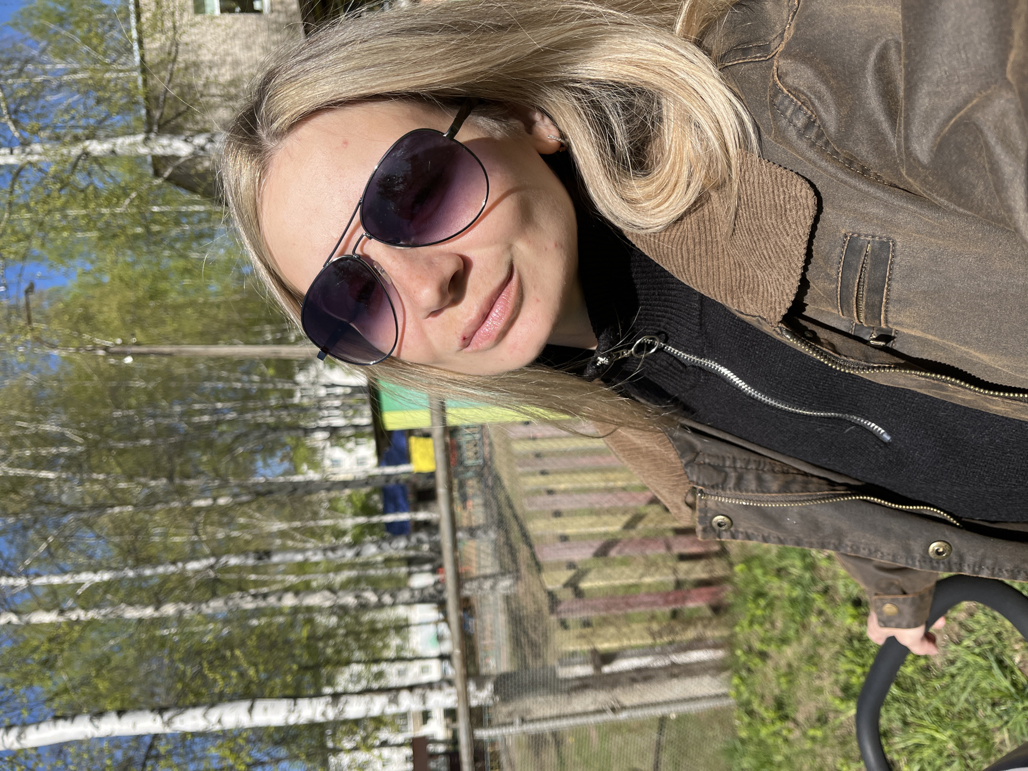 A blond woman wearing sunglasses and a brown jacket poses for a selfie in Chaikovsky, Russia. She is holding onto the handle of a black stroller, standing near birch trees and a colorful playground. Clothes hang on a line between apartment buildings in the background.