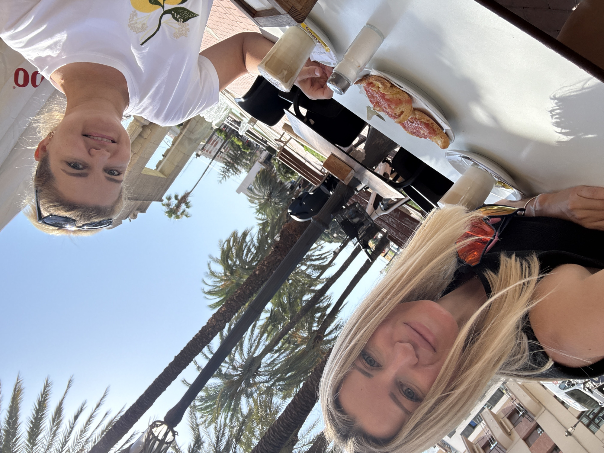 Two women are taking a selfie outdoors at a cafe. The woman in the foreground on the right is smiling and wearing a white t-shirt with a floral design and sunglasses on her head. The woman on the left, with blonde hair, is also smiling and wearing a black shirt with sunglasses tucked into the neckline. They are seated at a white table with a salt shaker, a glass of coffee, and two pastries with what appears to be tomato on them. In the background, palm trees, a fountain, and buildings suggest an urban setting, likely in a warm climate like Valencia, Spain, given the presence of palm trees and the style of architecture.