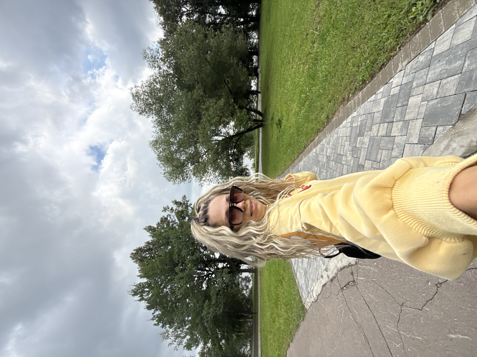 In Dumino, Russia, a woman with long, wavy blonde hair and sunglasses poses for a selfie in a park. She is wearing a yellow hooded sweatshirt with a red design on the front. The background features a paved walkway, a grassy area, and trees. The sky is overcast with clouds.