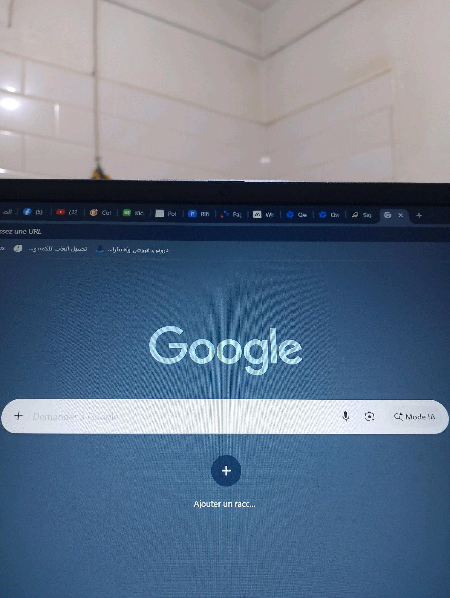 The image shows a close-up of a laptop screen displaying the Google homepage and several browser tabs. The laptop appears to be in an indoor setting, possibly a home or office, with white tiled walls visible in the background. The screen shows the Google logo and a search bar with the text "Demander à Google" (Ask Google). To the right of the search bar, there are icons for a microphone, Google Lens, and "Mode IA" (AI Mode). Below the search bar, a large plus icon is visible, with the text "Ajouter un racc..." (Add a shortcut...) underneath. The browser tabs at the top show various websites, including social media icons, a YouTube icon with a notification count, and tabs with Arabic text. The overall impression is of a functional digital workspace. The image does not contain any elements that place it specifically in Bordj Menaiel, Algeria, or any other specific geographical location.
