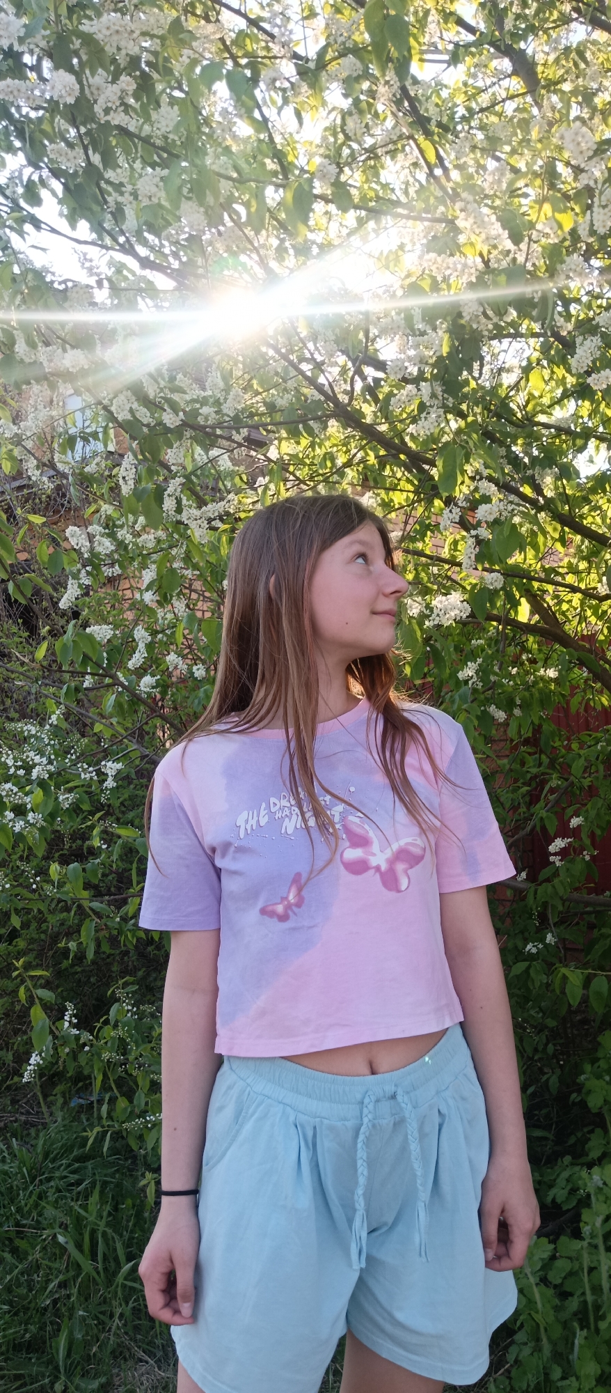 A young woman stands outdoors in Unknown, Unknown. She has long brown hair, and she is wearing a pink and purple tie-dye crop top with a butterfly graphic and light blue shorts. The background shows a tree with white flowers in bloom, partially obscuring the sun. The scene appears to be shot during daylight.
