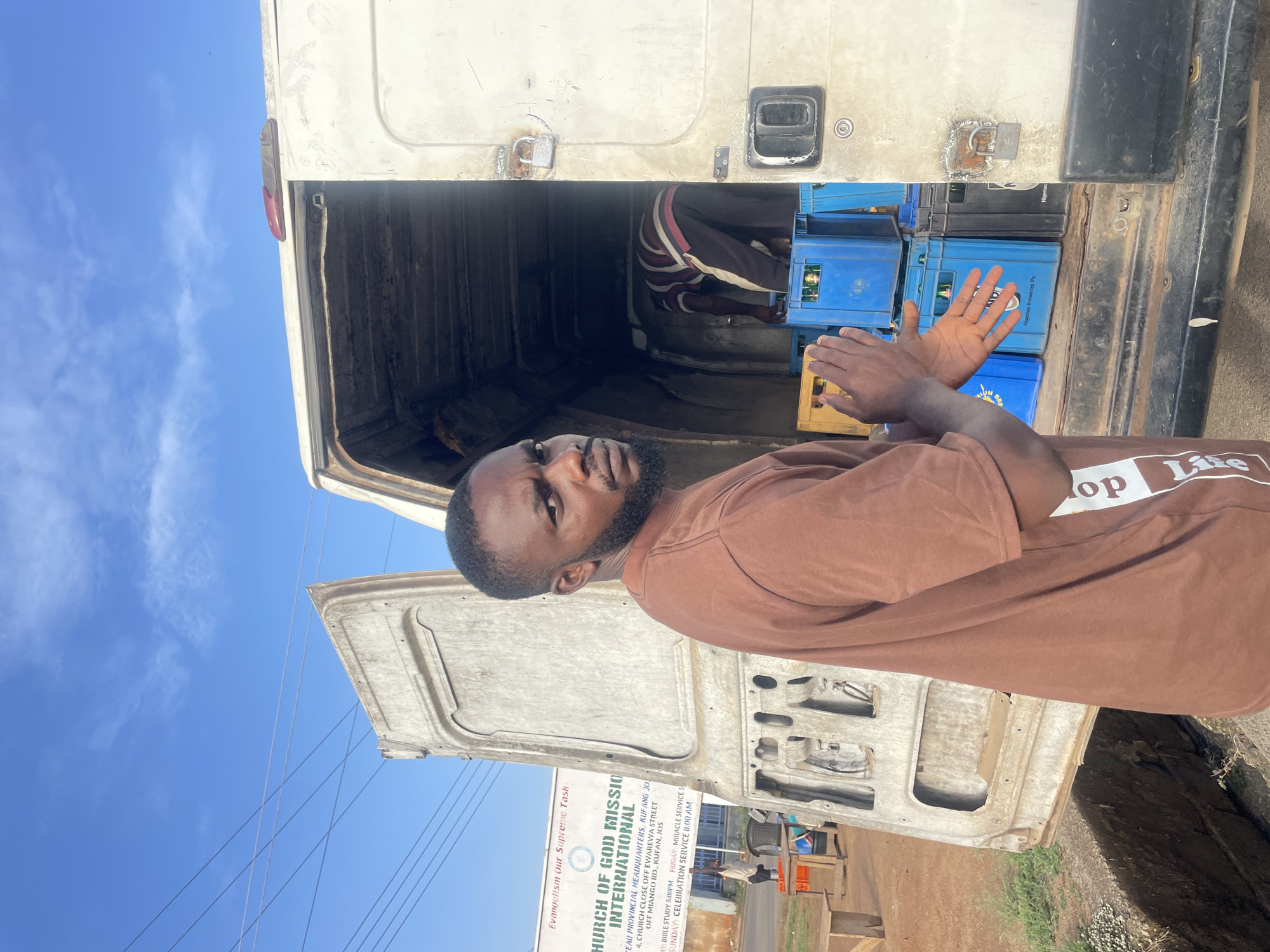A medium shot, taken at eye-level, shows a man unloading blue and yellow crates from the back of a white truck in Jos, Nigeria. The man is dark-skinned, with a beard, and wears a brown shirt with white lettering. He is turned to the side, facing the camera, and clapping his hands. Behind him, another man is visible in the truck. The crates appear to contain beer, possibly from Nigerian Breweries Plc. The truck's back doors are open, and a sign for "Church of God Mission International" is visible in the background, advertising services and bible study in the Kufang area of Jos. Wires are visible against a blue and cloudy sky.