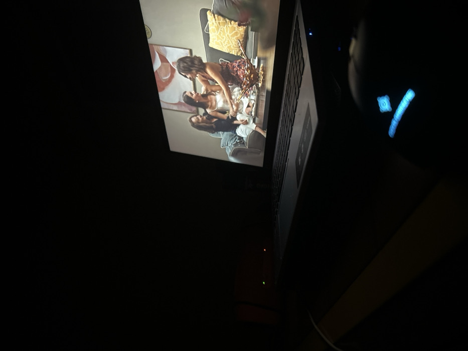 A laptop screen displays a scene of three women, likely from a movie or TV show, sitting on a sofa in a brightly lit room. The image is captured in a dark environment, with the laptop tilted slightly upwards. A mouse, with a blue LED light, is positioned to the right of the laptop. The overall composition is dark and centered on the laptop screen.
