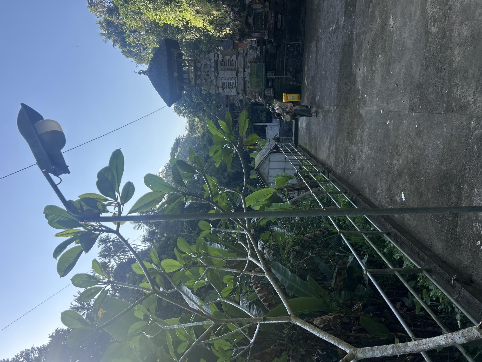 In the city of Bangli, Indonesia, a paved walkway leads towards a Balinese temple complex. A tall, dark-colored lamp post stands on the right side of the walkway, casting a shadow. Lush green foliage, including large leaves and thin branches, frames the left side of the image, with some trees in the background. The temple structure, with its multi-tiered thatched roof and intricate stone carvings, is the prominent subject in the background. Two individuals, one carrying a bag, are walking away from the camera towards the temple. A yellow trash bin is visible near the entrance to the temple. The sky is clear and blue, with sunlight filtering through the trees.