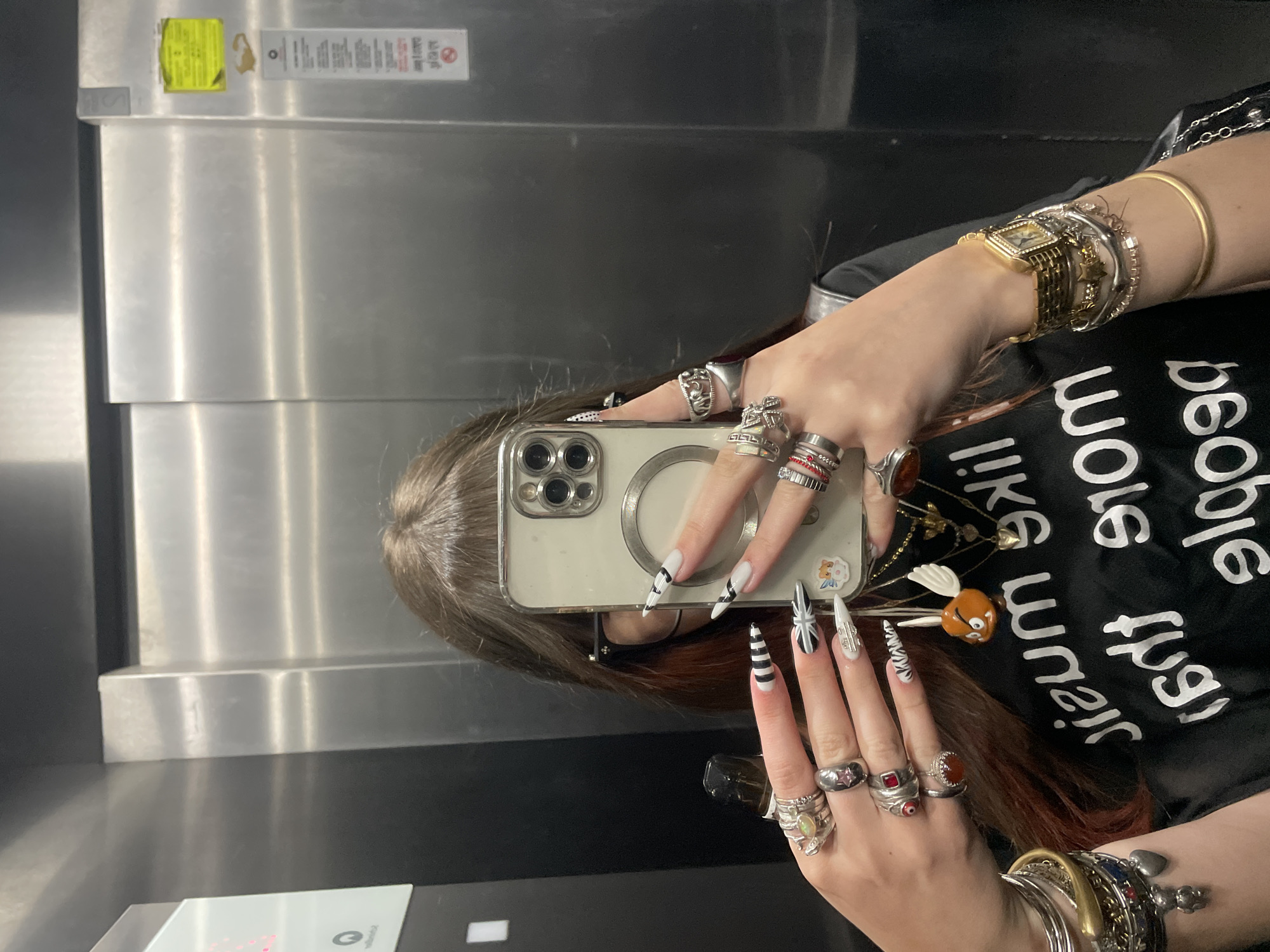 A person is taking a mirror selfie in what appears to be an elevator. The person has long, decorated nails and is wearing numerous rings and bracelets. The person is wearing a black shirt with white lettering. The elevator has stainless steel walls. There is no evidence in the image to indicate the location is Raanana, Israel.
