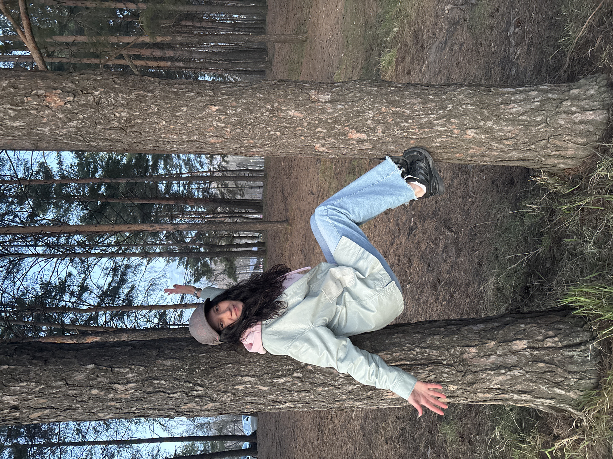 A young woman is depicted lying on a large, horizontal tree trunk in a forest setting, captured in a rotated perspective that makes the trunk appear to be suspended horizontally between vertical trees. She is smiling and looking towards the camera, with her body bent in a relaxed pose. She is wearing a light blue jacket over a light pink top, blue jeans that are ripped at the knee, black chunky sneakers, and a grey baseball cap. Her long, dark hair is visible beneath the cap.
Her left arm is extended upwards, while her right hand rests on the tree trunk near her waist. Her legs are bent, with her right foot placed on the trunk and her left foot slightly dangling. The tree trunk itself is thick and covered in rough, dark brown bark, consistent with a pine or conifer.
The background shows a dense forest of tall, slender trees, likely pines, with a light blue to grey sky visible through their branches. The ground is covered with dirt, sparse dry grass, and some moss, indicating a natural, unpaved environment. The overall lighting suggests it is either late afternoon, early evening, or an overcast day, giving the scene a soft, diffused quality. No specific weather conditions are discernible, but her attire suggests cool temperatures. There is no visible text in the image. The scene is set in a wooded area, with the provided context indicating it is in Chaikovsky, Russia.