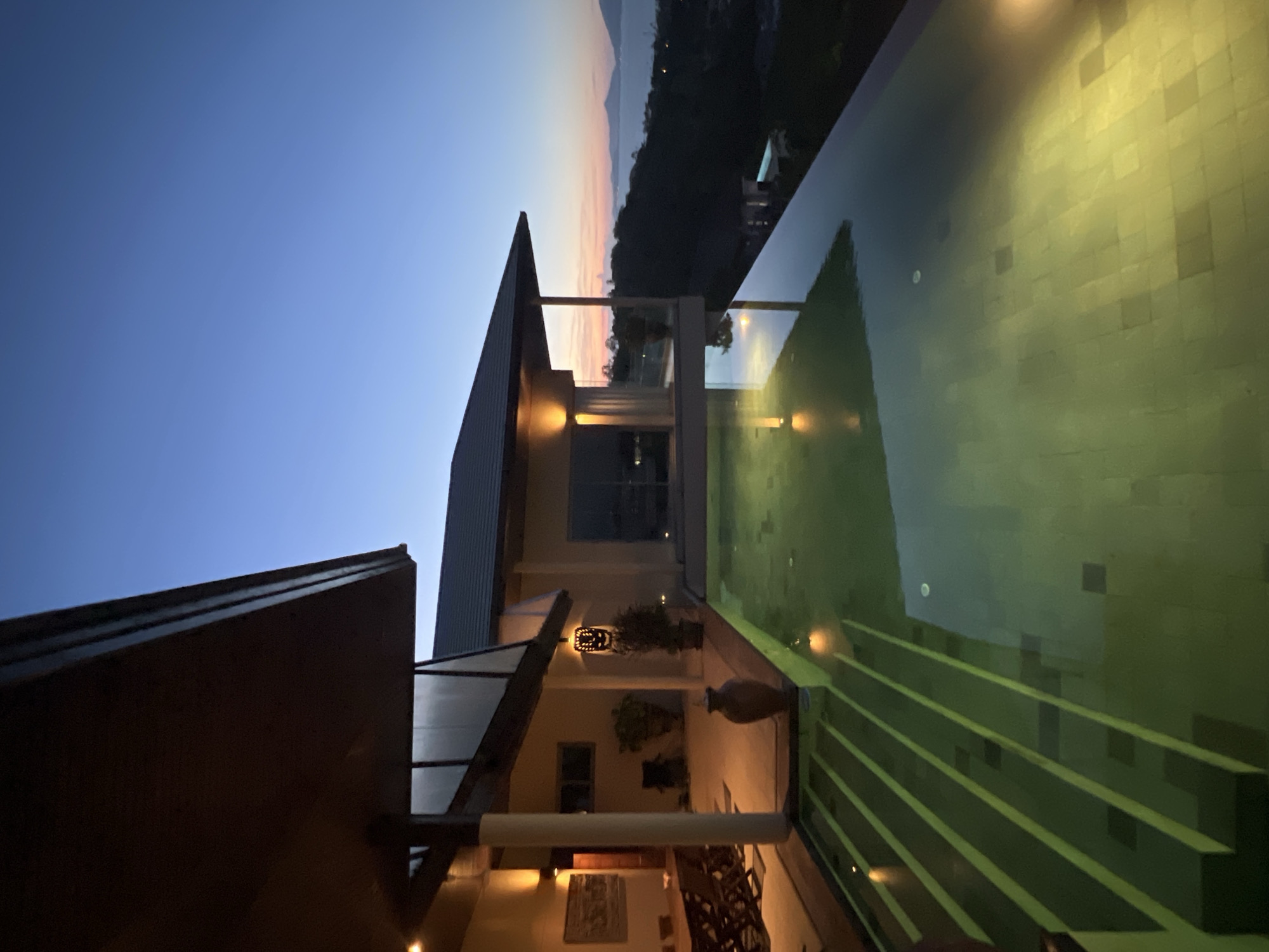 The image depicts an infinity pool at dusk in Baan Bo Phut, Thailand, overlooking a body of water and a hilly coastline. The pool's water is a greenish hue, illuminated by underwater lights, and reflects the ambient light and the structures around it. Steps are visible leading into the pool from the left side, with a few lights embedded within them. To the left of the pool is a covered patio area with a wooden dining table and chairs. A large, decorative urn sits near the edge of the patio. The architecture of the building is modern, with a dark, flat roof and exterior lighting. The sky transitions from a deep blue at the top to soft orange and pink hues near the horizon, indicating either sunrise or sunset. The overall scene conveys a sense of luxury and tranquility.