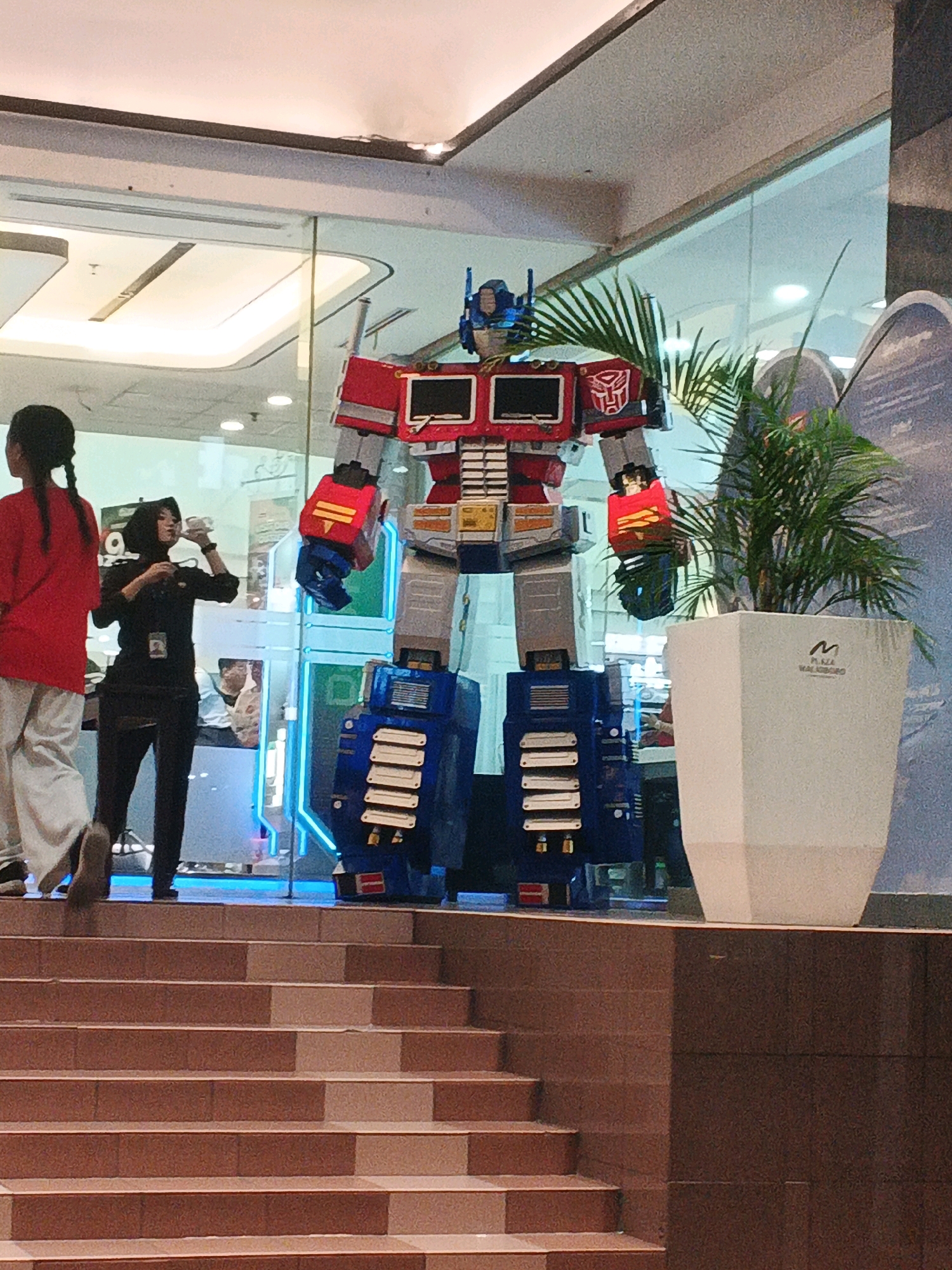 The image depicts a life-size Optimus Prime statue located inside a shopping mall, likely Plaza Malioboro in Yogyakarta, Indonesia, as suggested by the logo on the planter. The statue is a prominent feature, positioned near a large potted plant. In the foreground, a set of brown-tiled stairs leads up towards the statue. To the left of the statue, a person wearing a red shirt and white pants is partially visible, facing away from the camera and walking. Next to them, a woman in a black outfit, possibly a mall employee, is standing and holding a drink. The background shows a glass wall revealing interior elements of the mall, including lighting fixtures and other decorative items. The overall environment suggests a public indoor space, possibly a promotional display within the mall.