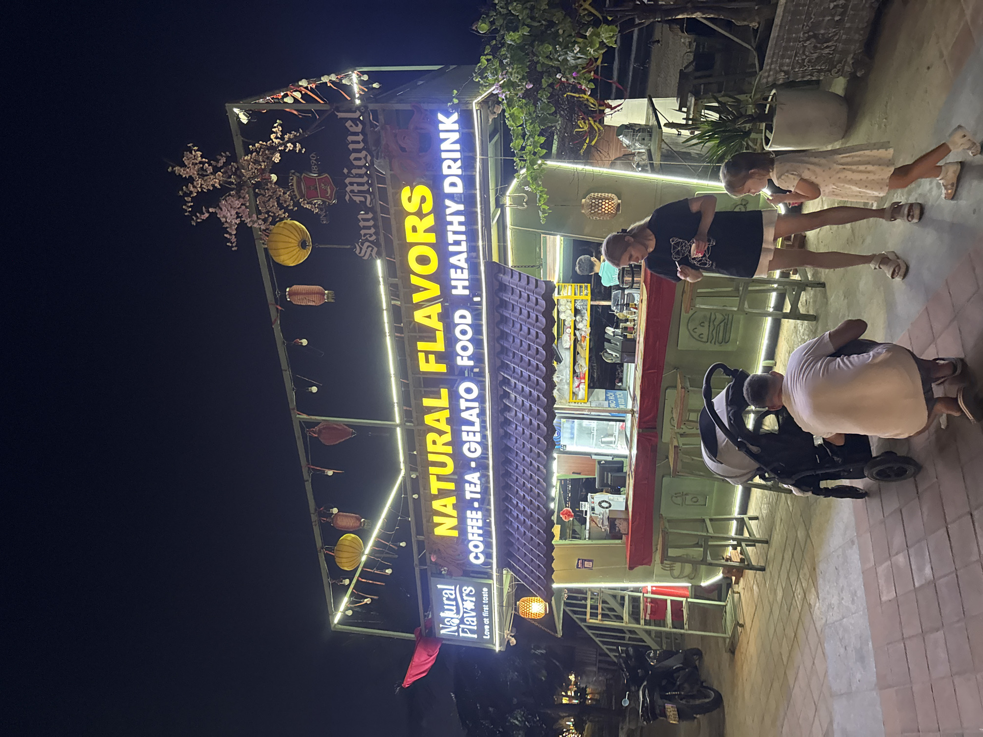 Captured at night, the image showcases the "Natural Flavors" establishment, located in Đà Nẵng, Vietnam. The business is brightly lit with a sign displaying "Natural Flavors" in yellow letters above "Coffee-Tea-Gelato Food Healthy Drink." The sign also features smaller lettering indicating "San Miguel" and the phrase "Love at first taste." Decorative lanterns are hung along the sign, adding to the ambiance. People are visible in the foreground. A woman stands next to a child, both seemingly enjoying a treat. A man bends over a stroller. The establishment's layout reveals tables and chairs for patrons. A motorcycle is parked nearby, suggesting local transport. The surrounding environment indicates an urban setting.