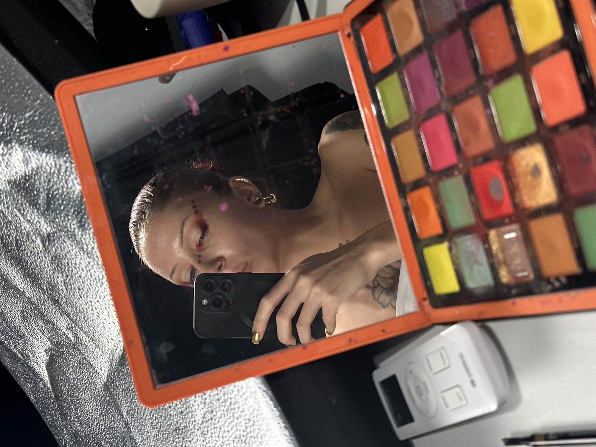 The image displays a mirror selfie, featuring a person, likely a woman, reflected in an orange-framed compact mirror. The other side of the compact holds a colorful eyeshadow palette with numerous pans of varying shades including reds, oranges, greens, yellows, purples, and glittery tones, many showing signs of use and some smudging.
The reflection shows the woman with light-colored hair, pulled back, with noticeable pink streaks. She wears prominent reddish-pink makeup around her eyes and small matching dots on her forehead and cheek. A nose ring and a gold-colored hoop earring are visible. Tattoos adorn her arm and shoulder. She holds a black smartphone with a triple-lens camera system, her hand adorned with gold nail polish, reflecting the act of taking the picture.
The background, visible both directly and in the reflection, includes a textured silver material on the left, possibly a reflector or fabric. On the right, a white electronic device with buttons and a screen, along with a dark pen-like object, is visible. The mirror's surface itself is marked with pink smudges, consistent with makeup application. The scene depicts a moment of self-documentation, highlighting an artistic or bold makeup look. The image is set in North Kuta, Indonesia.