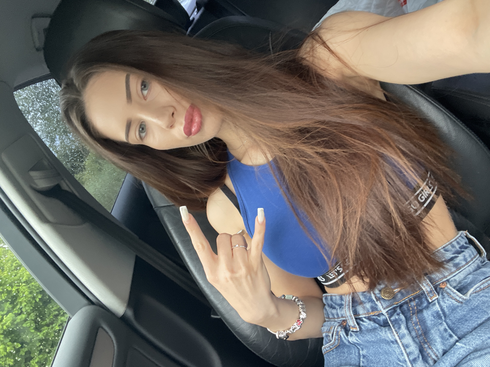 A young woman with long brown hair and blue eyes is sitting in the passenger seat of a car. She is wearing a blue crop top and blue jeans, and she is making a rock-on hand gesture. The car appears to be parked, with green trees visible through the windows. The weather seems to be rainy, as evidenced by water droplets on the window.