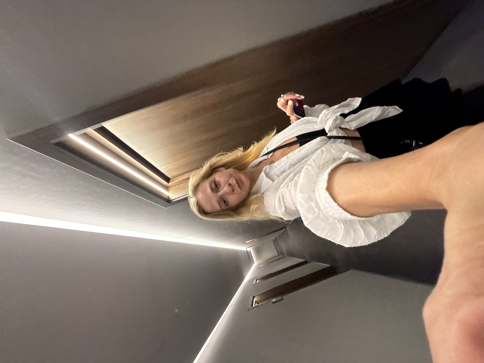 A woman with blonde hair is standing in a dimly lit hotel corridor, holding a phone in her outstretched hand, taking a selfie. She is wearing a white open shirt tied at the waist over a black top, and black pants. The corridor features several closed doors on either side, illuminated by linear lighting along the ceiling. The location is identified as a hotel corridor in Kerch, Ukraine.