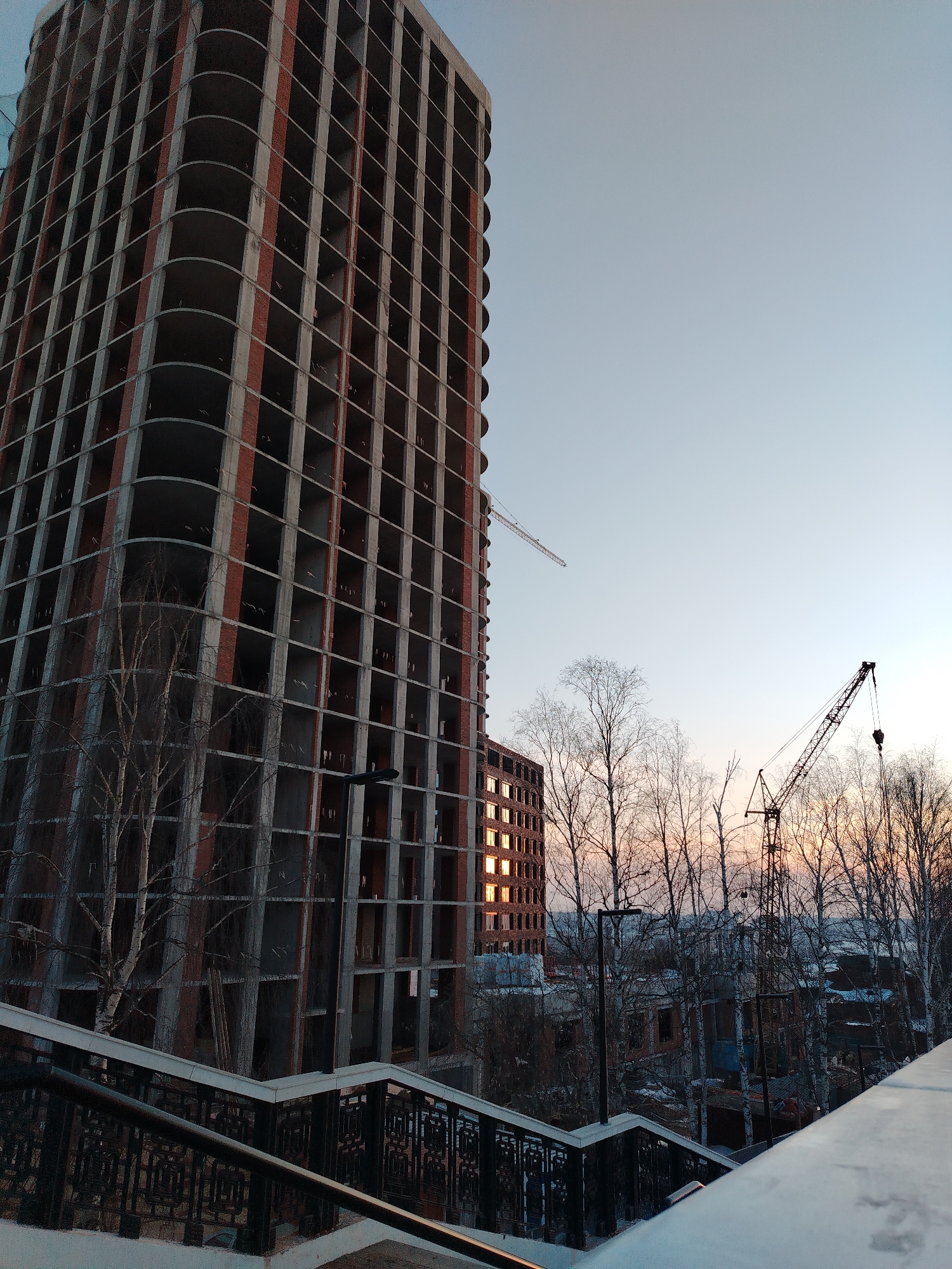 The scene depicts an urban landscape under a twilight sky, identified as being in Izhevsk, Russia. In the left foreground, a set of outdoor stairs with dark, ornate metal railings descends, partially covered in snow or frost. The primary subject is a tall, multi-story building under construction, dominating the left and central frame. This building features an exposed concrete framework with vertical sections of red brickwork and a distinct rounded corner, appearing to be an unfinished high-rise. A tower crane is visible near the top of this structure. To the right of the main building, another mid-rise structure, also appearing to be under construction or renovation, has several windows illuminated by a low-angle sun, suggesting either sunrise or sunset. A second, larger mobile crane is positioned further back, silhouetted against the horizon. Numerous bare deciduous trees with thin branches, likely birches due to visible lighter bark, are scattered across the midground. The sky transitions from a light blue overhead to shades of orange and pink near the horizon. No people are visible within the frame.