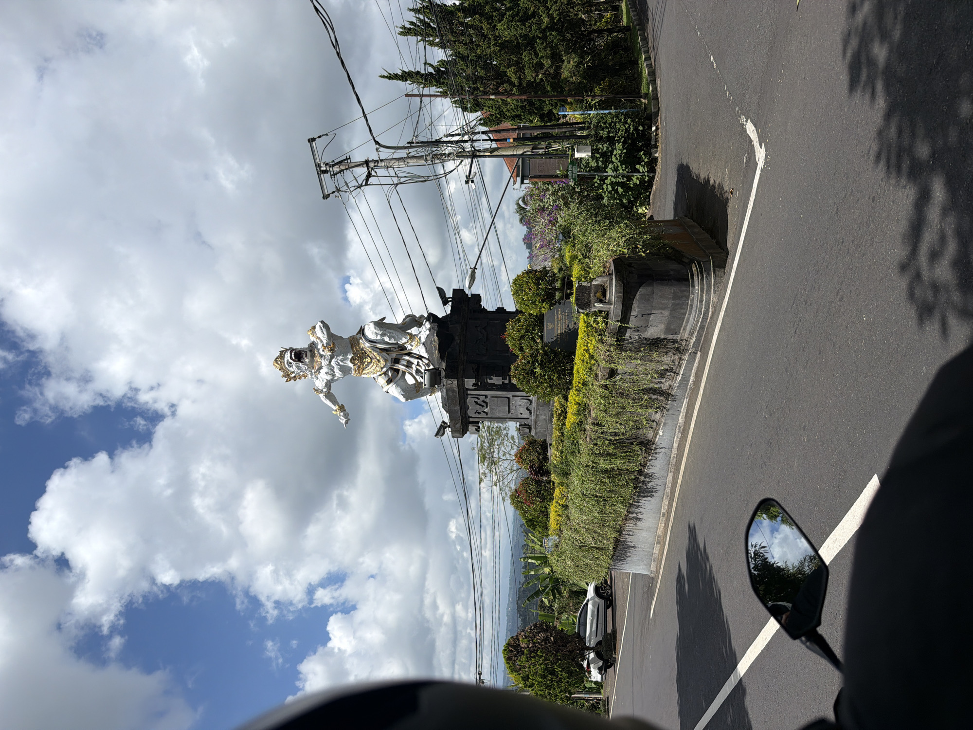 The image captures a statue of a Balinese deity, likely a representation of Hanuman, a significant figure in Hinduism known for his strength and devotion. The statue is white with gold accents and is positioned on a dark pedestal adorned with intricate carvings. The deity is depicted in a powerful stance, with its mouth open and arms outstretched, conveying a sense of action or a dramatic moment.
The statue is situated in a prominent location, likely a roundabout or a public square, in the town of Petang, Indonesia. It is surrounded by lush greenery, including well-maintained hedges and flowering bushes, which are characteristic of tropical landscapes. Behind the statue, a white car is parked on the side of the road, and further in the background, a dense forest and rolling hills can be seen, suggesting a rural or semi-rural setting within Petang.
The sky is a mix of blue with scattered white clouds, indicating a bright day. Electrical poles and wires are visible, suggesting that this is a developed area with infrastructure. The foreground of the image shows a section of a paved road with white lane markings. The reflection in the motorcycle's rearview mirror reveals more of the sky and some trees, adding depth to the scene. The overall impression is that of a vibrant and culturally rich environment.