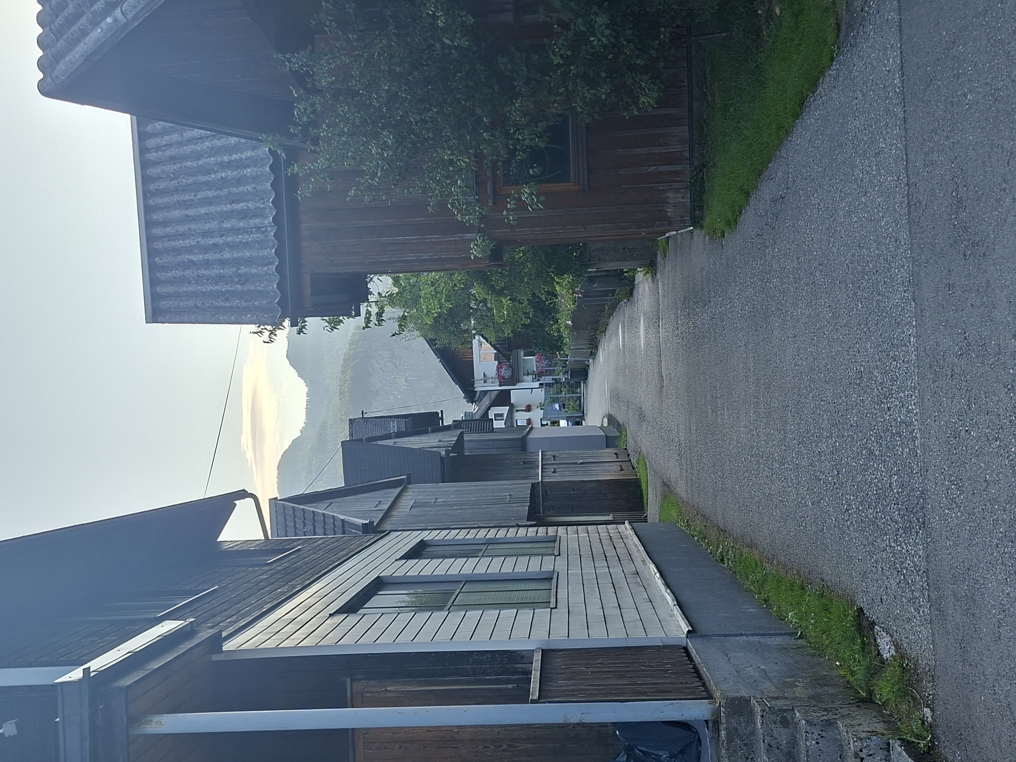 The image depicts a narrow street in Hallstatt, Austria, lined with traditional wooden houses. The houses feature dark wood siding and steep roofs, some covered with corrugated material. The street is paved with asphalt and appears to be a pedestrian walkway or a narrow residential road. On the left side of the street, a light-colored house with horizontal siding and a covered porch is visible, with concrete steps leading up to it. On the right side, a dark wooden house with a large window and dense green foliage obscuring part of its facade is prominent. The street recedes into the distance, leading towards a backdrop of gentle, tree-covered mountains under a hazy sky. The overall scene conveys a quiet, rustic atmosphere typical of a European alpine village. No people are visible in the image.