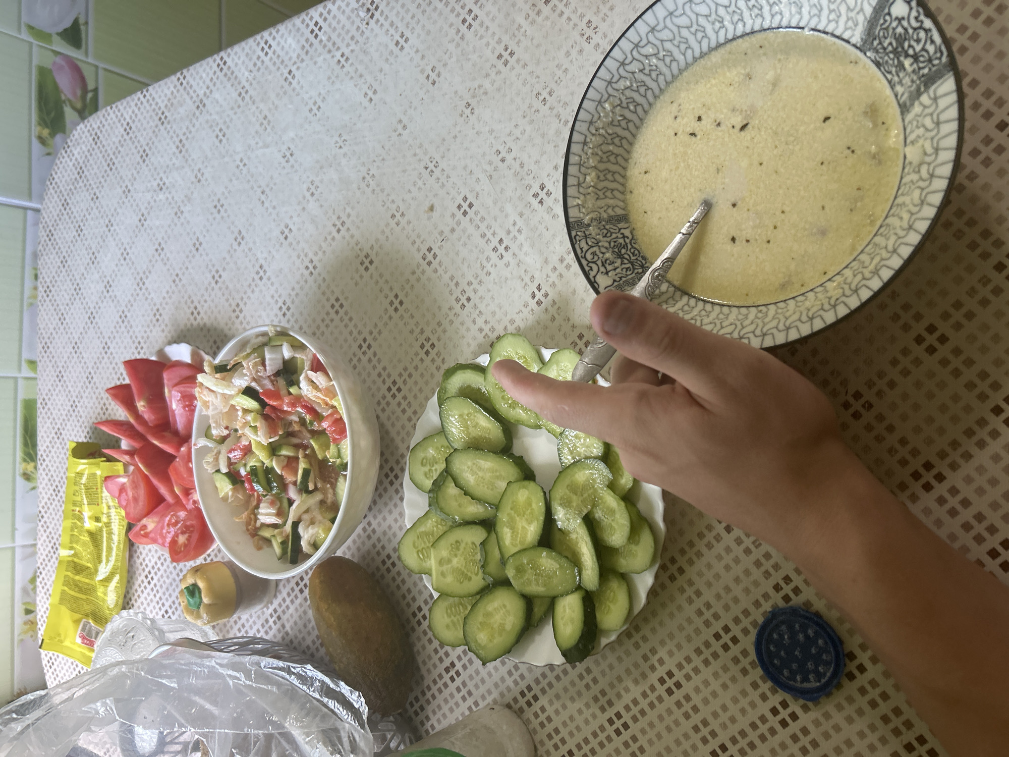 A close-up, overhead shot shows a meal laid out on a checkered tablecloth. In the foreground, a person's hand with a silver spoon is reaching into a bowl of pale yellow soup with visible flecks of herbs and possibly small pieces of food. To the left of the soup bowl is a plate filled with sliced cucumbers. Behind the cucumbers is a whole pickle. Further back, a bowl contains a salad made of chopped tomatoes, cucumbers, and possibly cabbage or lettuce, with a light dressing. To the left of the salad bowl is a decorative toothpick holder shaped like a pumpkin. A yellow package is also visible behind the tomatoes, which are arranged in wedges on a white plate. A plastic bag and other items are partially visible in the upper left corner. The background shows a tiled wall in a kitchen setting. The overall scene depicts a simple home-cooked meal, potentially in a rural Russian kitchen, given the mention of Старотумбагушево.
