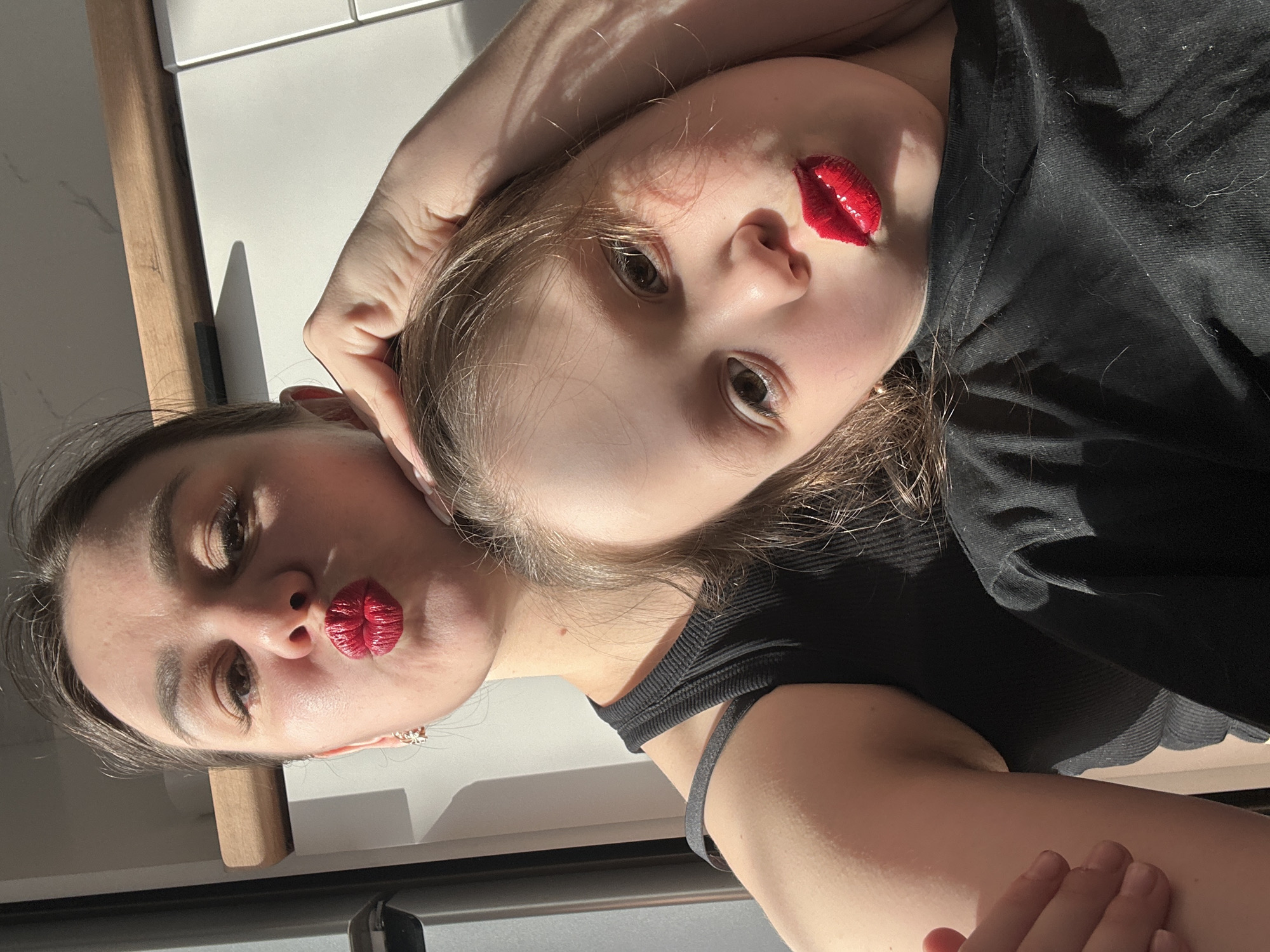 A selfie image captures a woman and a child, both facing the camera with puckered lips accentuated with red lipstick. The child is positioned at an angle above the woman. The woman is wearing earrings and the child is wearing a black top. The setting appears to be indoors, with sunlight casting shadows on a white surface behind them. The specific location is not discernible from the image.
