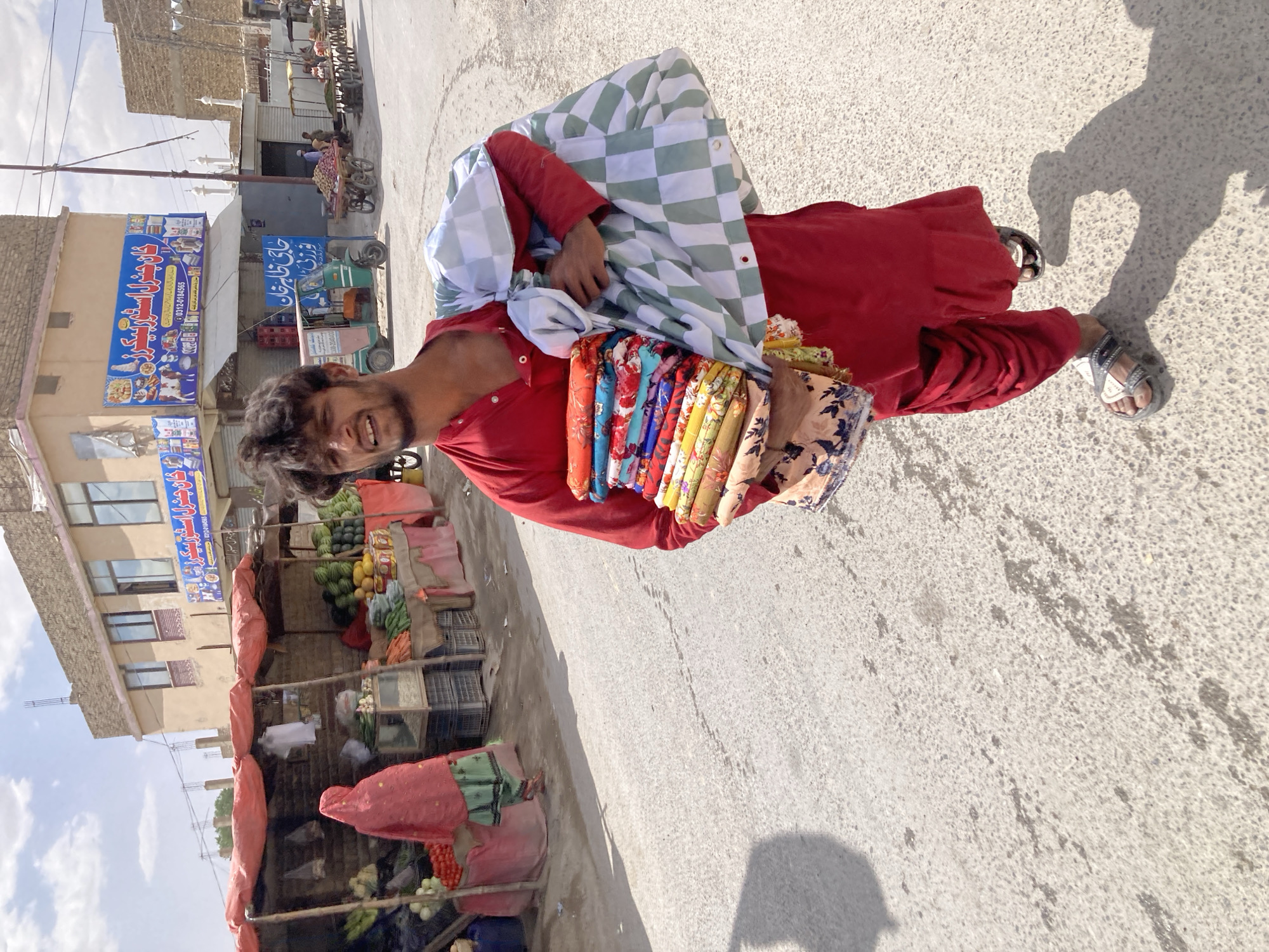 The image captures a vibrant street scene, likely a market, in Quetta City Tehsil, Pakistan, under a bright, sunny sky. Strong shadows on the ground indicate a midday or late morning setting.
The main subject is a man standing on a paved street, smiling broadly at the camera. He wears a red shirt and matching red trousers, along with sandals. Draped over his left arm and shoulder is a white and green checkered cloth, while his right arm holds a bundle of neatly folded, colorful textiles with various patterns, suggesting he is a street vendor.
To the left, a bustling market lines the street with several makeshift stalls. These stalls are covered with red or orange tarps and display a variety of fresh produce, including watermelons, bananas, apples, onions, tomatoes, and other vegetables in crates and baskets. People are visible near these stalls, engaged in daily activities. One individual in red and green clothing is partially visible behind a vegetable stand.
Buildings of varying construction, some appearing rustic with exposed brickwork and others with glass windows, form the backdrop. On one prominent building, a large blue sign in Urdu reads "چوہدری اسٹیورز" (Chaudhry Stores) with "خوش آمدید" (Welcome) and a phone number (0312-0141565). Below it, another partially visible blue sign has "حاجی طالبان" (Haji Talib...) or similar text. An auto-rickshaw is parked near these shops, and further down the street, carts, possibly laden with goods, can be seen. The overall scene depicts a lively market environment.