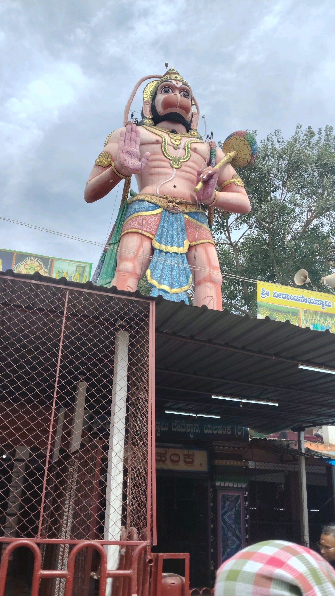 The image shows a large statue of Hanuman, a Hindu deity, standing on top of a structure. The statue is painted in bright colors, with a pinkish-orange skin tone, blue and gold clothing, and a golden headdress. Hanuman is depicted with his left hand raised in a gesture of blessing or protection, and his right hand holding a mace. In the background, there are trees and a cloudy sky. Below the statue, there is a building with a corrugated metal roof. There is a wire fence in the foreground, and a person wearing a checkered headscarf is visible at the bottom right of the image. The text on the yellow banner above the building appears to be in the Kannada language, which is spoken in the Indian state of Karnataka, including Bengaluru. Specifically, the text on the banner appears to refer to "Shree Veeranjaneya Swami," a common name for Hanuman. This suggests the location is a temple dedicated to Hanuman in or near Bengaluru.