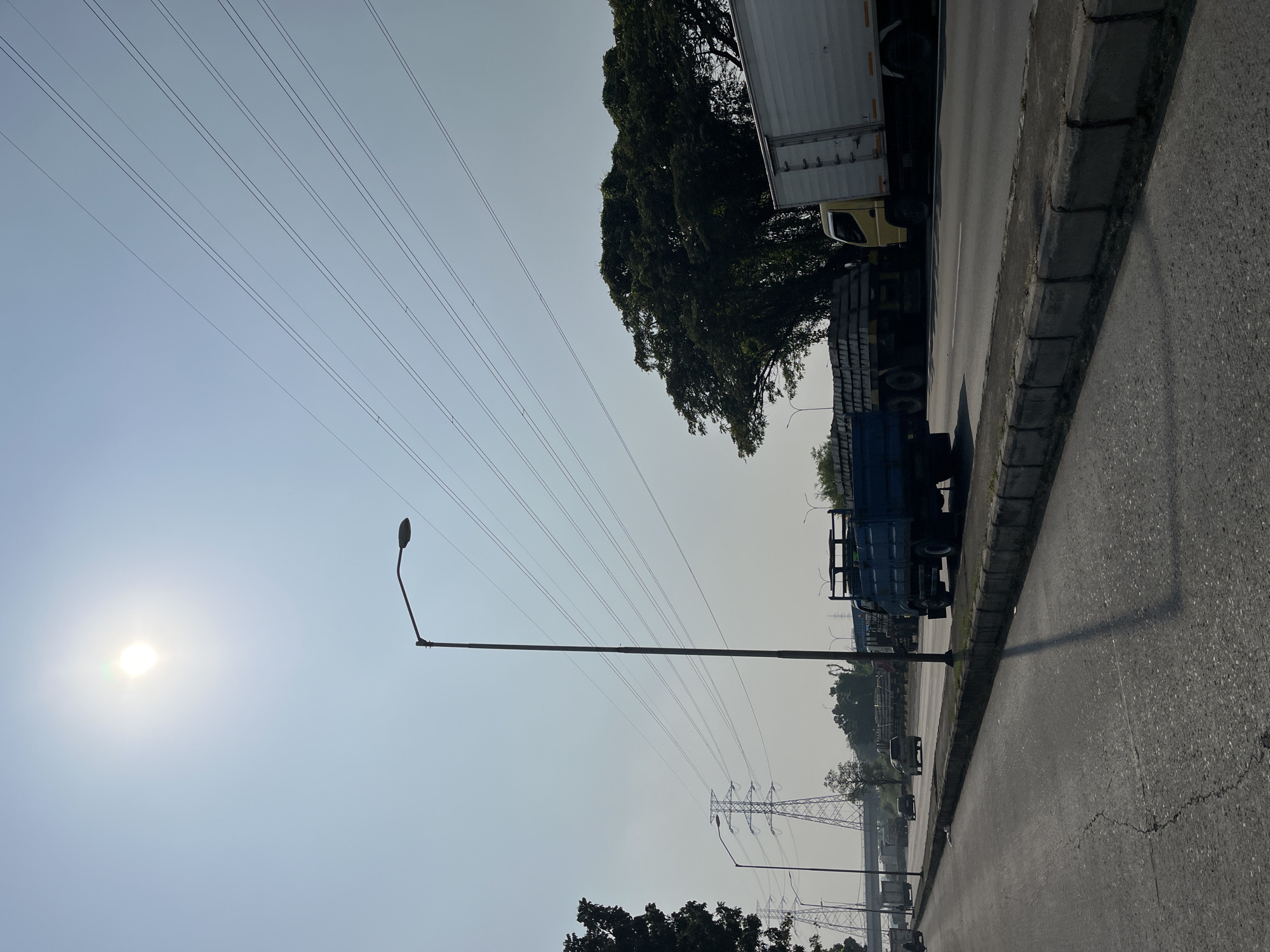The image depicts a sunlit road scene in the city of Wunut, Indonesia. In the foreground, a cracked asphalt road stretches into the distance, bordered by a concrete curb. A tall street lamp stands prominently on the right side of the road, casting a long shadow. In the background, several large trucks are visible on the road. One truck in the center is blue and appears to be a flatbed. To its right, a yellow and white box truck is parked. Further down the road, more trucks and vehicles are present, along with power lines stretching across the sky and electricity pylons in the distance. Lush green trees line the right side of the road. The sky is clear and bright, with the sun visible in the upper left quadrant of the image.