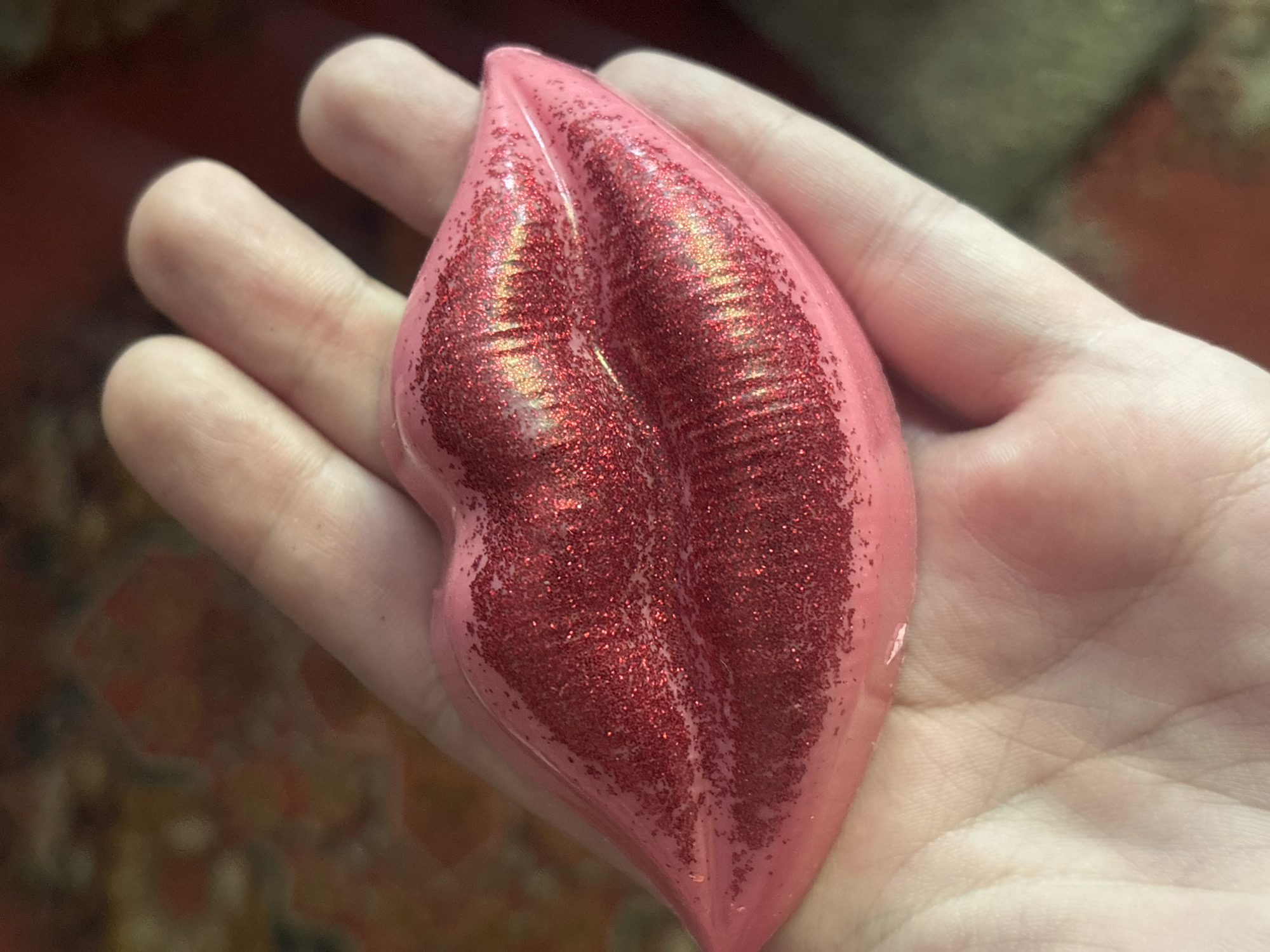 A close-up, overhead shot shows a person's hand holding a pink, lip-shaped object with red glitter. The object is placed in the center of the palm, with the fingers curved around it. The background is blurred, but appears to be a patterned surface.