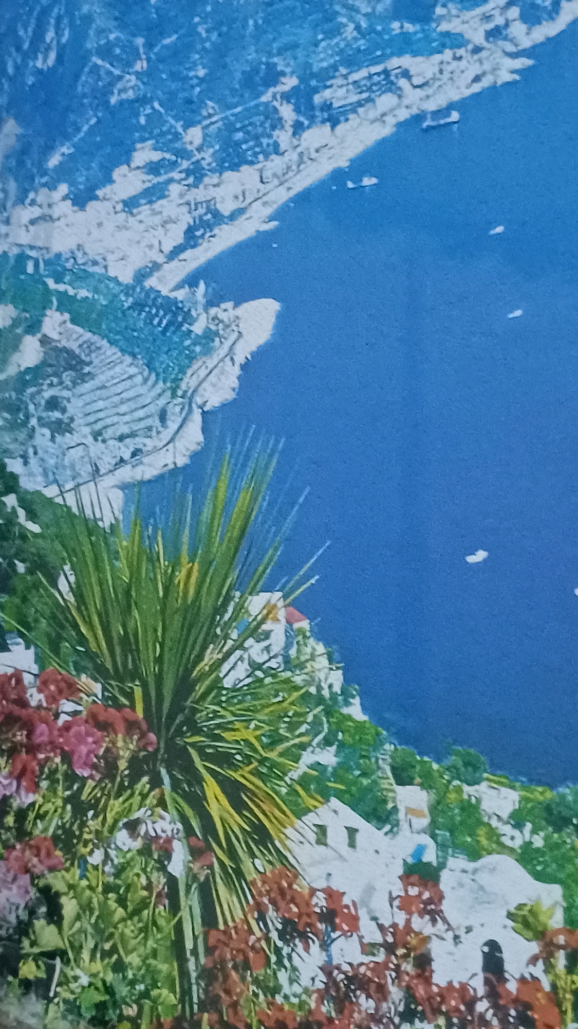 This image depicts a scenic coastal view with a prominent foreground of lush greenery and flowers. In the background, a body of blue water is visible, dotted with several small white boats. Along the coastline, a town or village with white buildings is nestled on a hillside, with terraced agricultural fields or structures on the steeper slopes. The overall impression is of a Mediterranean or tropical coastal landscape. The image does not contain any people or visible actions. No specific location within Wonokerso, Indonesia can be identified from the image.