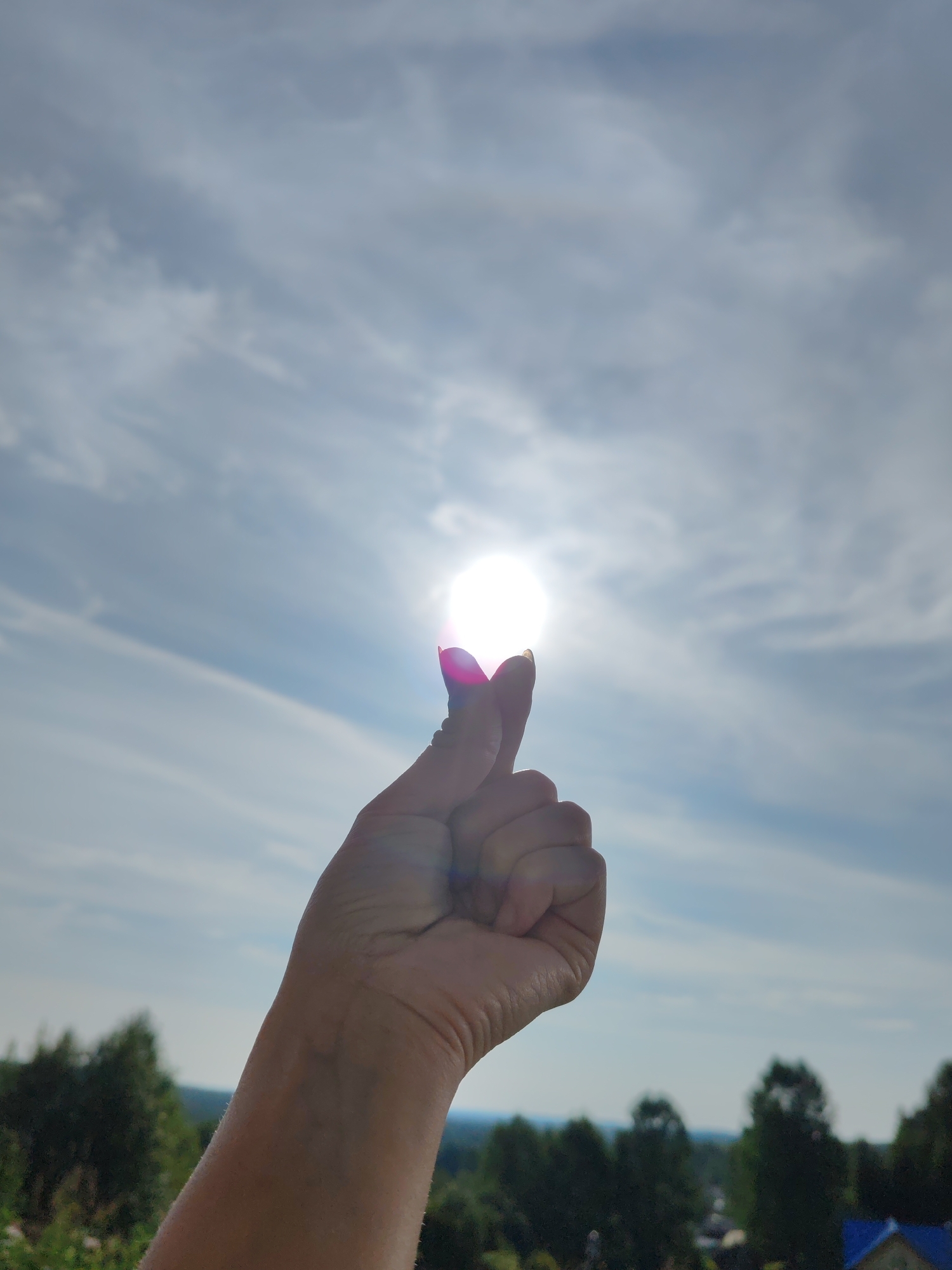 A close-up, low-angle shot captures a human hand silhouetted against a bright sky. The sun, a brilliant white orb, is positioned directly in the center of the frame, partially obscured by the fingertips of the hand, creating a lens flare effect with pink and purple hues. The hand, with a manicured nail on the index finger, is raised, as if trying to hold or block the sun. The sky is partly cloudy, with streaks of white clouds against a pale blue backdrop. In the background, out of focus, a landscape unfolds, featuring a dense line of dark green trees. Beneath the trees, hints of buildings with blue rooftops suggest a rural or suburban settlement, possibly within the city of Чухлэм, Russia. The overall impression is one of natural beauty and human interaction with the environment.