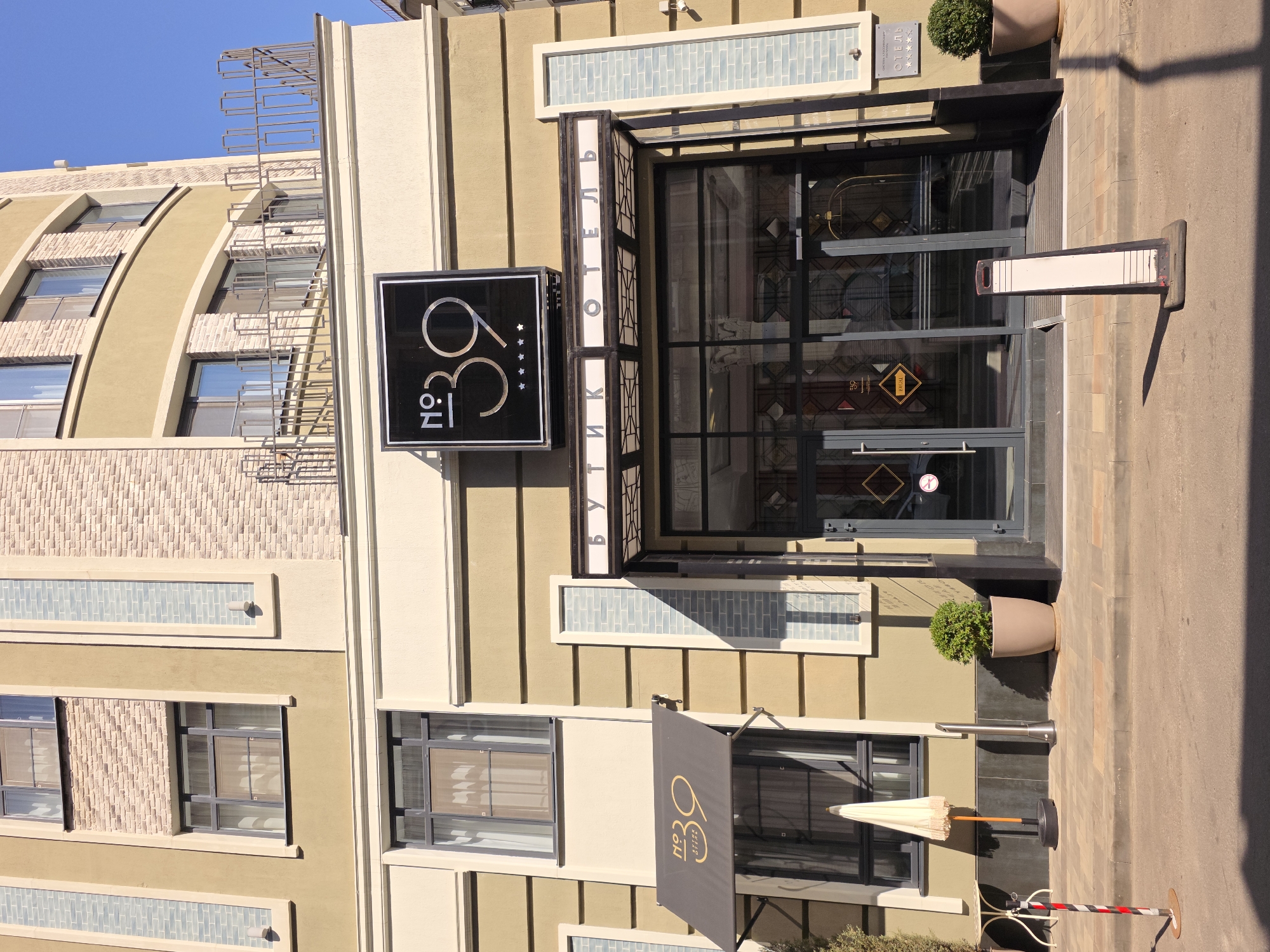 The image shows the entrance to the "No. 39 Boutique Hotel" in Rostov-on-Don, Russia. The hotel has a modern, European-inspired architectural style with light beige and olive-green facade elements, interspersed with vertical decorative panels of light blue brickwork. A prominent black sign with "No. 39" and five stars is displayed above the entrance. The entrance itself features large glass doors and a canopy with the hotel's name written in Cyrillic letters, "Бутик Отель". To the left of the entrance, a dark grey awning with "No. 39" and "Boutique Hotel" is visible, shading a window. In front of the hotel, there is a paved sidewalk with a small round table and an umbrella to the left, and a potted plant to the right of the entrance. A metal informational stand with a blank white sign is positioned on the pavement in front of the doors. The sky is clear and blue, indicating a sunny day.