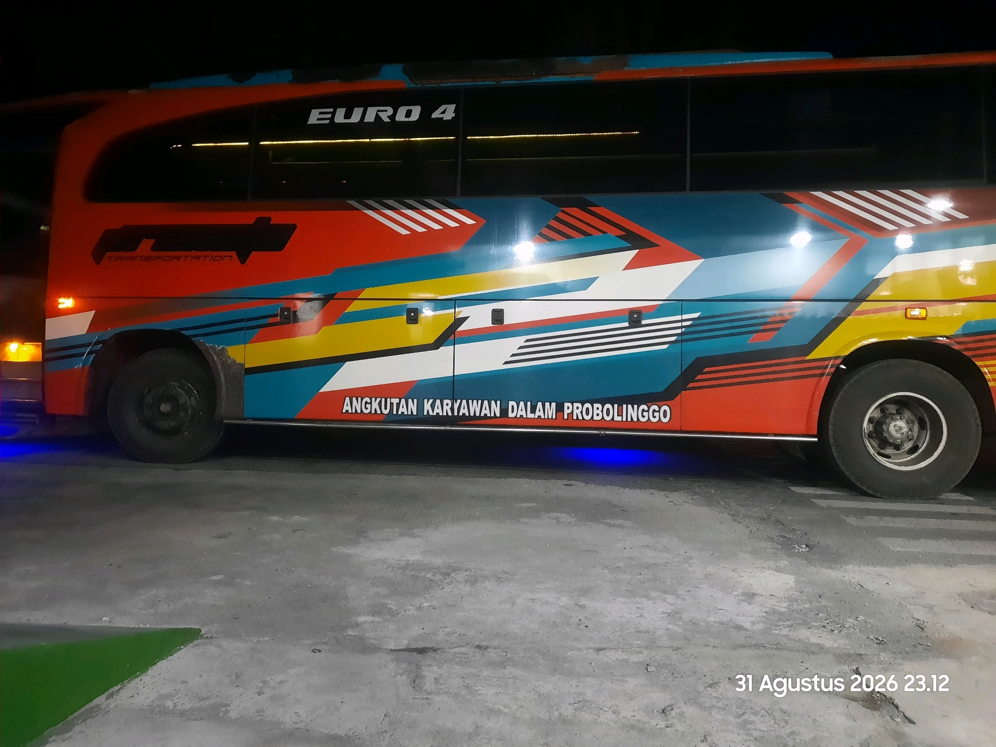 The image shows the side of a bus at night, parked on a concrete surface. The bus has a vibrant, graphic design in red, blue, yellow, and white. The text "EURO 4" is visible on the upper part of the bus. Below the windows, the text "ANGKUTAN KARYAWAN DALAM PROBOLINGGO" is written in white, indicating it is a staff transport service for Probolinggo. There is also a logo with the text "TRANSPORTATION" on the front section of the bus. The bus has two visible wheels, one in the front and one in the rear. Blue lighting is visible underneath the bus. The background is dark, suggesting it is nighttime. The ground is made of concrete, with some green painted markings visible in the lower left corner. A timestamp "31 Agustus 2026 23.12" is present in the lower right corner of the image. The image is taken from a slightly low angle, looking at the side of the bus. The location in Gending, Indonesia is inferred from the text on the bus, as Probolinggo is a regency in East Java, Indonesia, and Gending is a district within that regency.