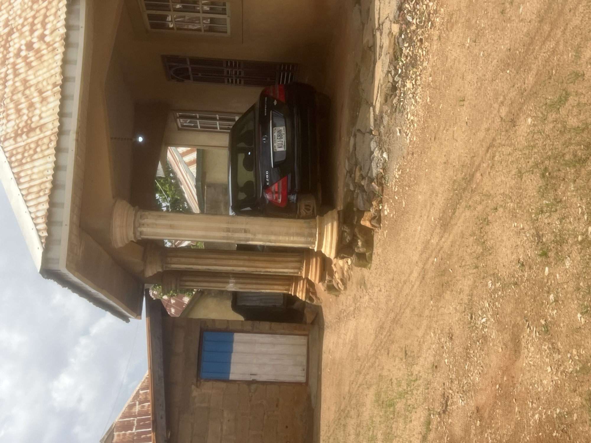 In Gura Top, Nigeria, a black Mercedes-Benz E200 sedan with license plate KUJ-68HN is parked on a dirt and gravel driveway in front of a building. The car is positioned under a covered porch supported by four fluted columns. To the left of the car is a brick building with a blue and white door. The main building to the right has a corrugated metal roof and windows with white frames. The sky is cloudy. The environment appears to be a residential area.