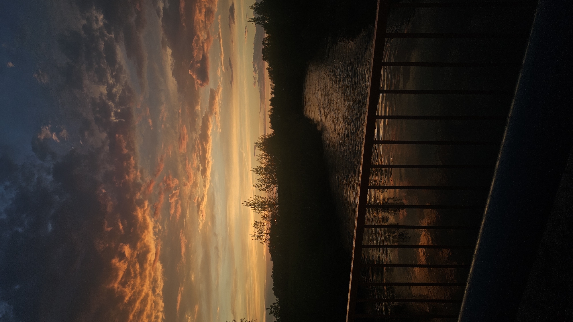 From an elevated vantage point in Yangiskain, Russia, a water body, possibly a river, reflecting the sunset, flows horizontally across the image. Silhouetted trees line the far bank. The sky features orange and yellow clouds against a gradient of blue. A railing runs along the right edge of the image.