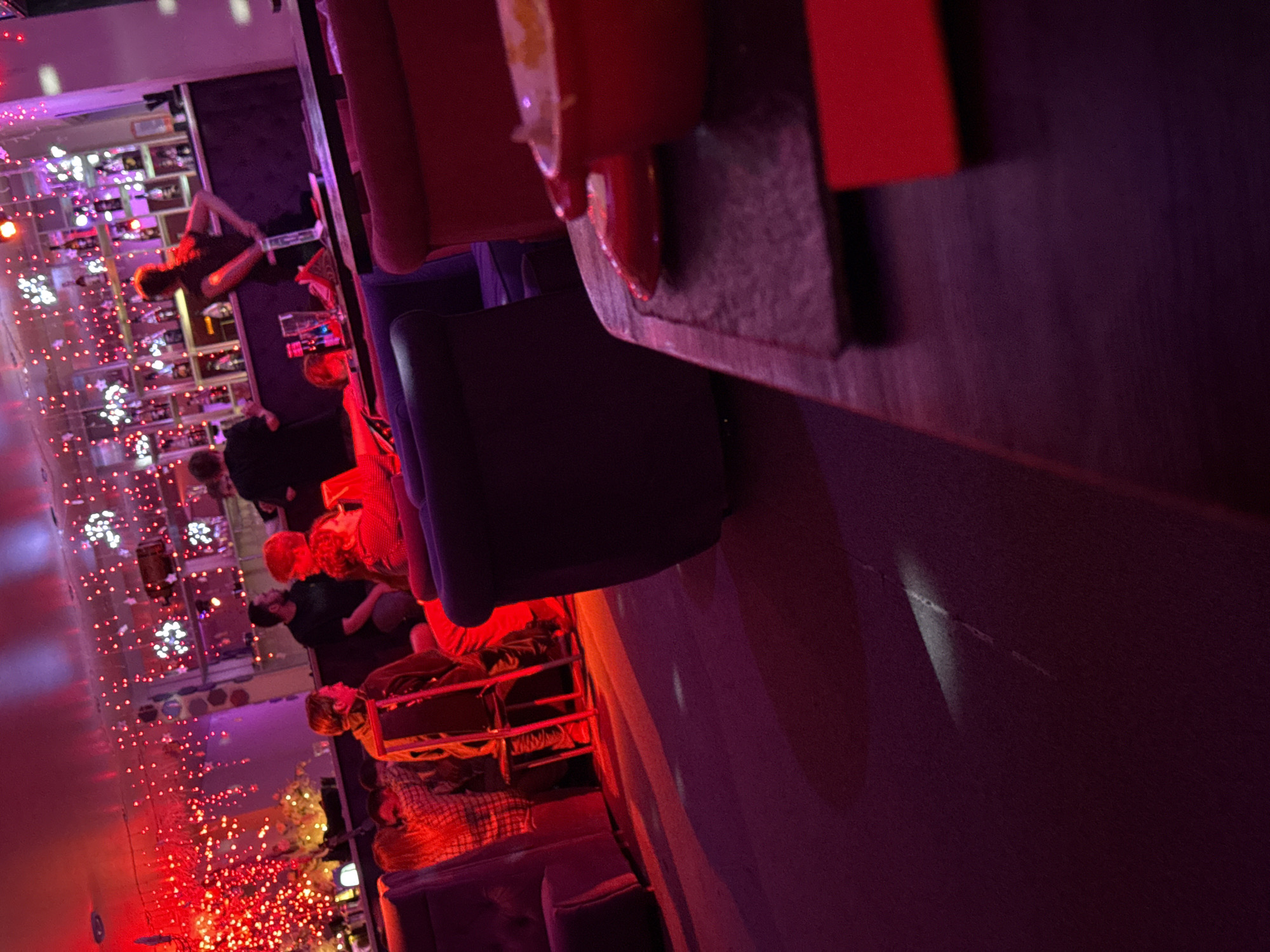 The image shows an interior space with several people seated and standing. A bar with lighted shelves and bottles is visible in the background, decorated with strings of red and white lights. Some individuals appear to be interacting with the bartender. Furniture includes chairs and sofas. The overall lighting is low, with red and purple hues dominating the color scheme. There is no information about the location in the city of Votkinsk, Russia.