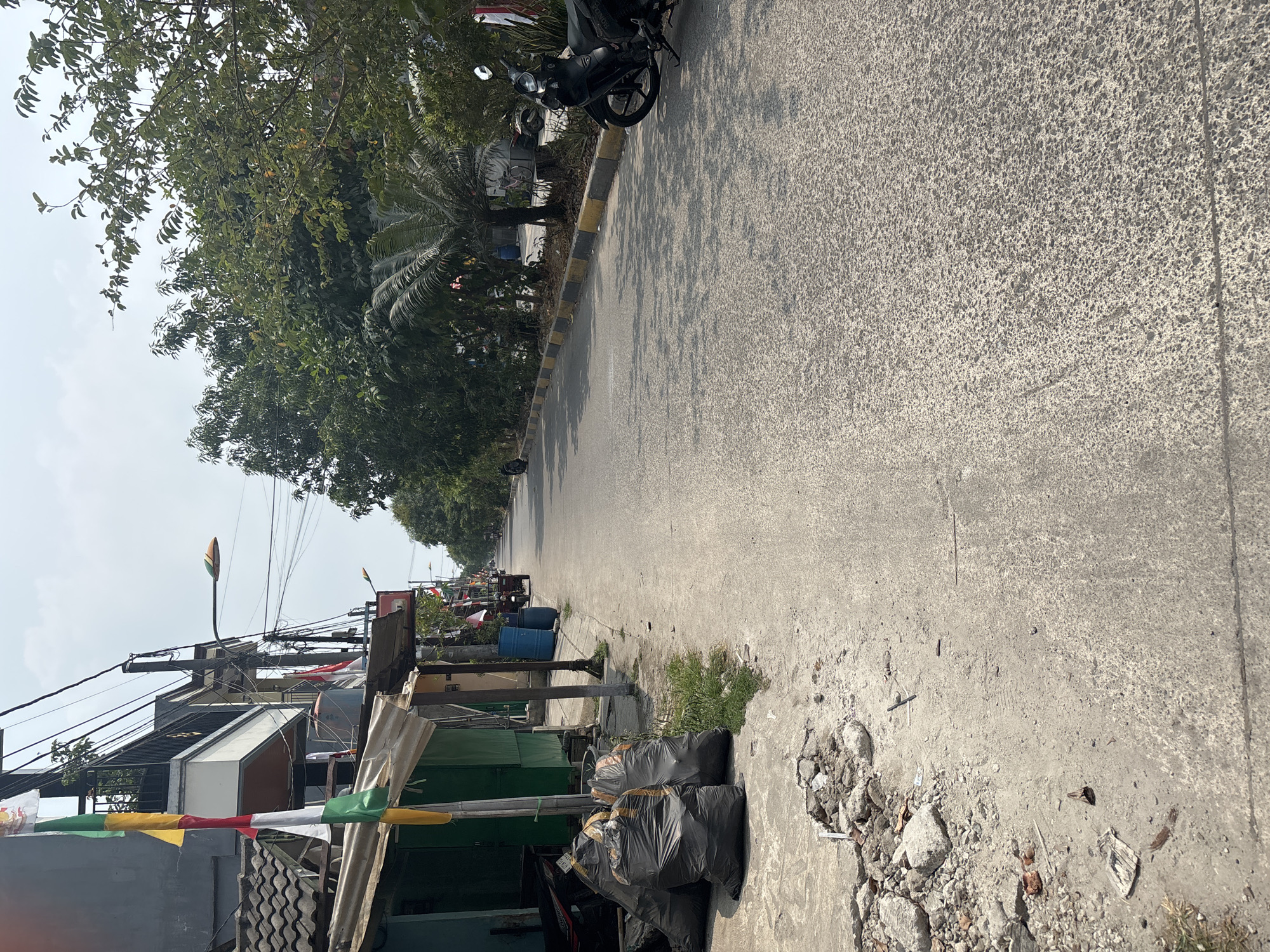 The image depicts a street scene in Telagamurni, Indonesia. On the left side of the street, a row of buildings with green facades and corrugated metal roofs are visible. Several large black trash bags are piled on the concrete sidewalk in front of these buildings. A motorcycle is parked to the left of the trash bags. In front of the buildings, there are poles with flags.
The street itself is paved with asphalt and curves to the right into the distance. The right side of the street is lined with trees and vegetation, with a curb marked by alternating yellow and black stripes. A black motorcycle is parked on the right side of the road, facing away from the camera. In the background, more trees and buildings are visible, as well as utility poles and wires spanning across the street. The sky is overcast. The overall impression is of a residential or commercial street in a tropical or subtropical climate.