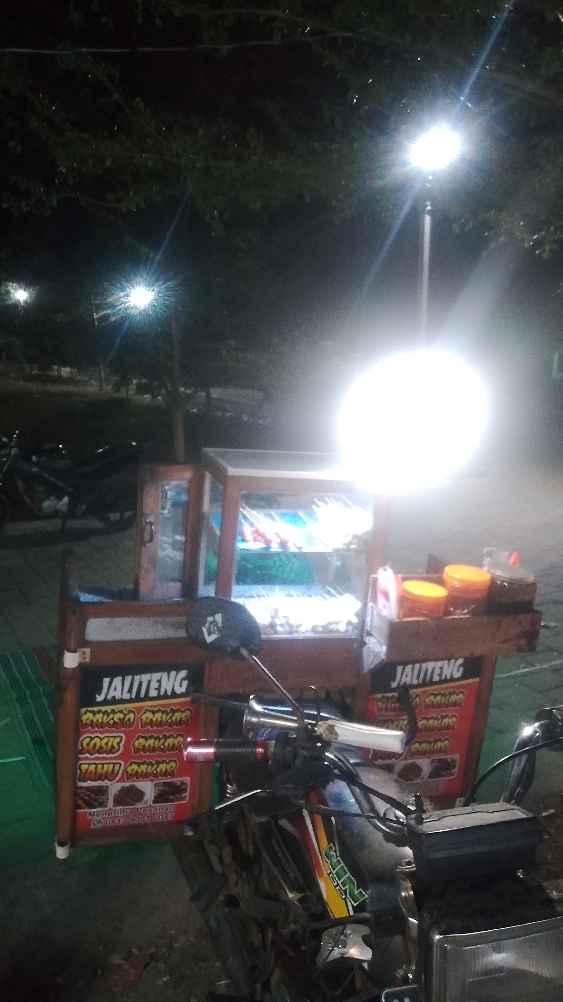 The image depicts a street food cart at night in Karanglangu, Indonesia. The cart, likely a mobile vendor, is illuminated by a bright overhead light and is attached to the front of a motorcycle. The cart itself is made of wood and glass, displaying rows of skewered food items, possibly satay or grilled meatballs. Various condiments in jars and plastic containers are also visible on a shelf attached to the cart. A sign on the cart reads "JALITENG" and lists "BAKSO BAKAR," "SOSIS BAKAR," and "TAHU BAKAR," indicating grilled meatballs, sausages, and tofu are for sale. Below this, it states "Menerima Pesanan" (Accepting Orders) and provides a phone number: "0813 9081 0087." The background shows trees and more streetlights, suggesting an outdoor, possibly public, setting within the city. The overall scene captures a typical nocturnal street food vending operation in Indonesia.