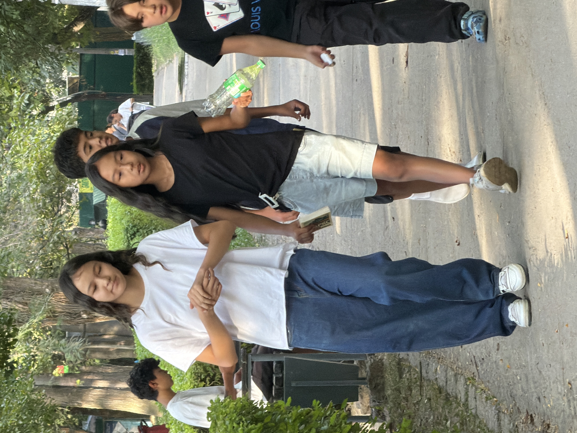 In an outdoor urban setting, a group of young people walk along a paved path bordered by green foliage and trees. In the foreground, a girl with long dark hair and wearing a white t-shirt and wide-leg blue jeans walks from left to right, her hands clasped in front of her. To her right, another girl with dark hair wears a black t-shirt and light-colored denim shorts, carrying a plastic bottle in her right hand. Behind them, a boy with dark hair and a grey jacket walks, and further to the right, a boy in a black t-shirt with a graphic print walks, holding a white object in his hand. In the background, other individuals and greenery are visible.