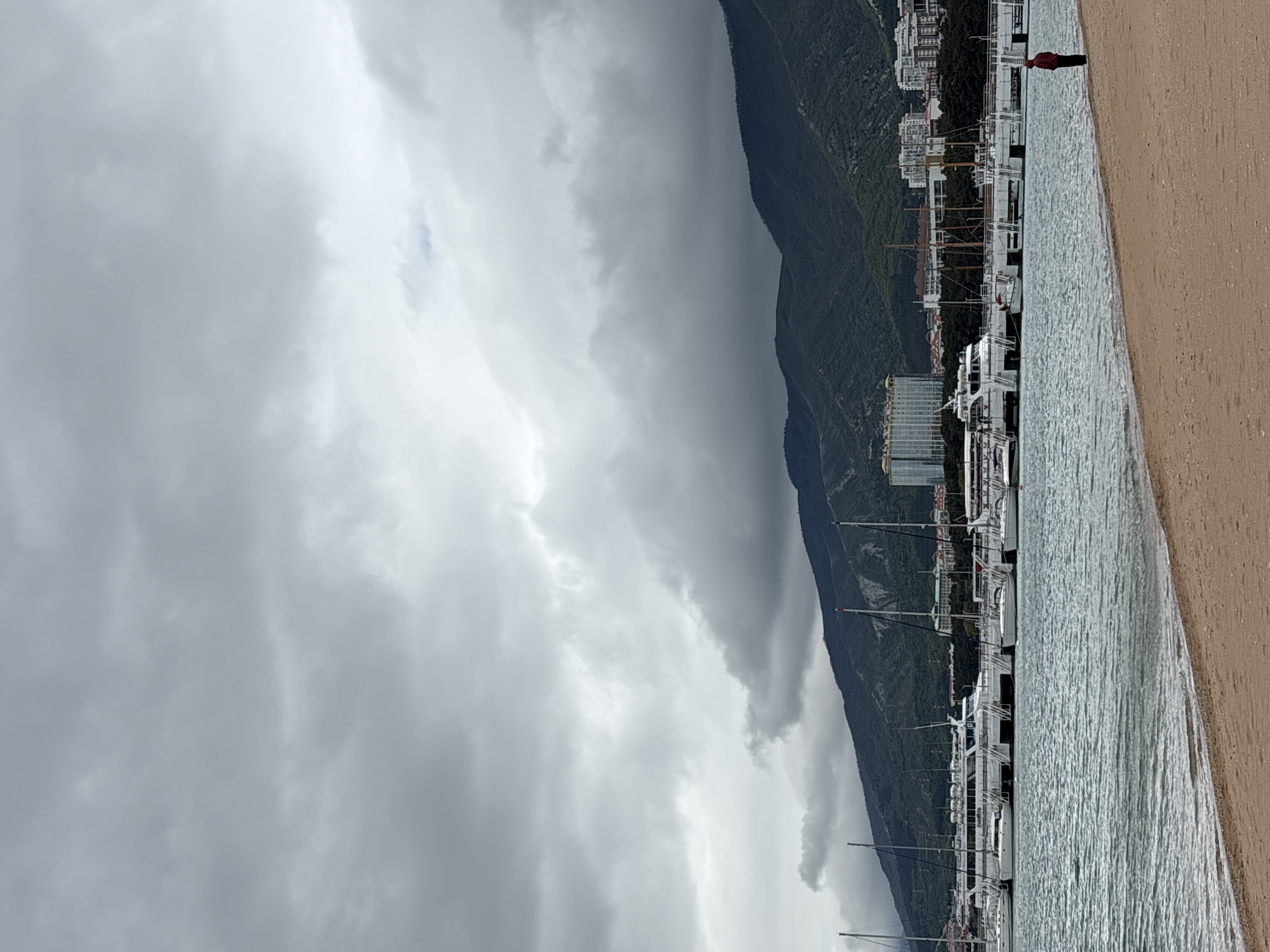 A scenic view in Gelendzhik, Russia, features a beach on the right side of the frame with a single person in the upper right corner standing on the edge of the sand, appearing to gaze out towards the sea. Adjacent to the beach is a harbor with numerous boats docked, extending parallel to the shoreline. Behind the harbor, a green, mountainous landscape rises up, reaching towards a sky dominated by heavy, overcast clouds, which suggest an impending storm. Buildings are also visible clustered behind the docks.